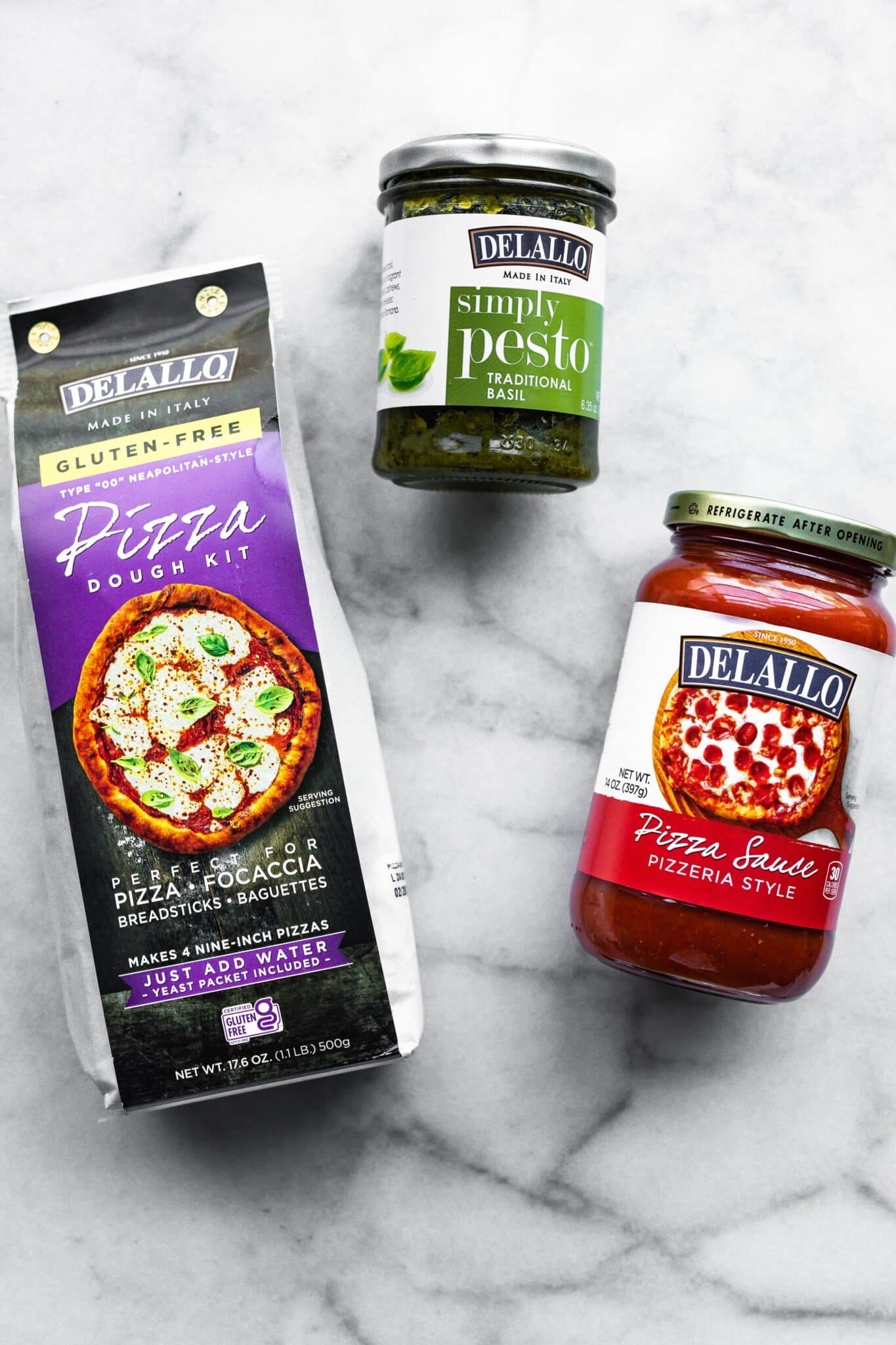 Pizza dough mix, pesto, and Delallo pizza sauce on a marble countertop.