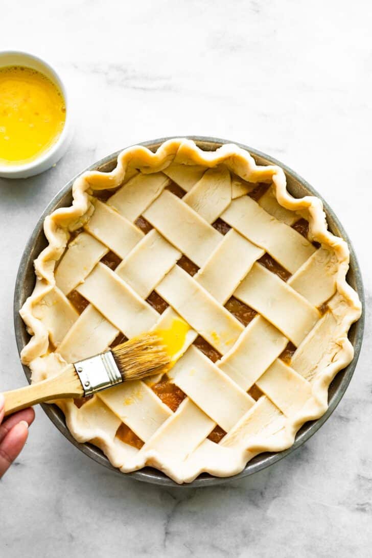 woman's hand brushing butter on an unbaked apricot pie with a lattice crust