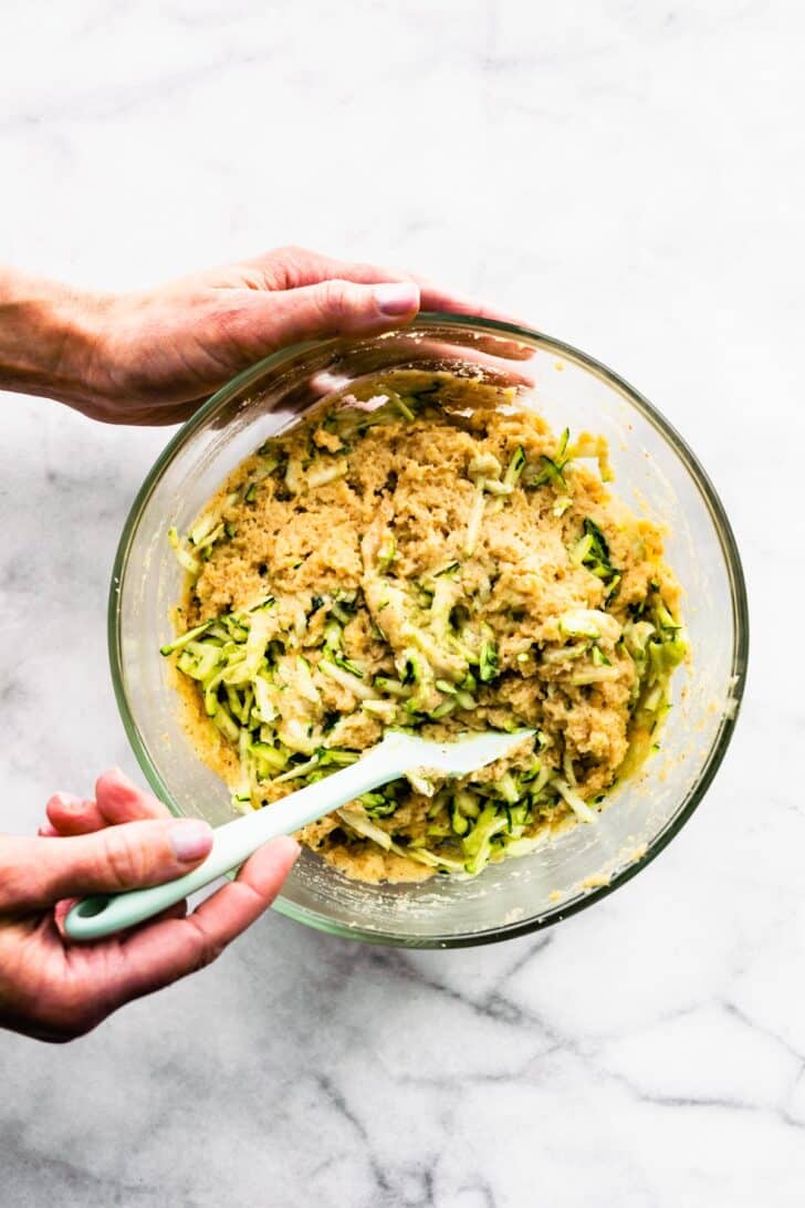 Two hands stirring shredded zucchini into bread batter.
