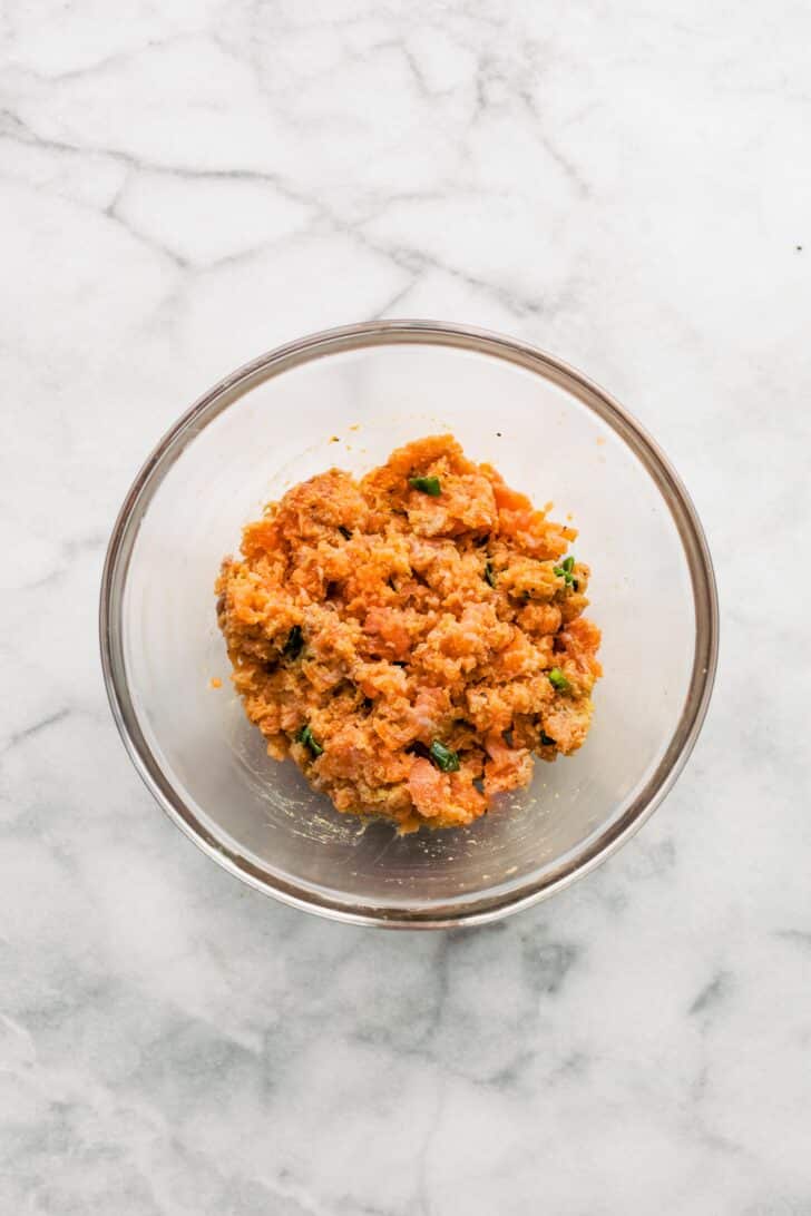 Chopped salmon mixed with binders like panko and veggies in a glass bowl.