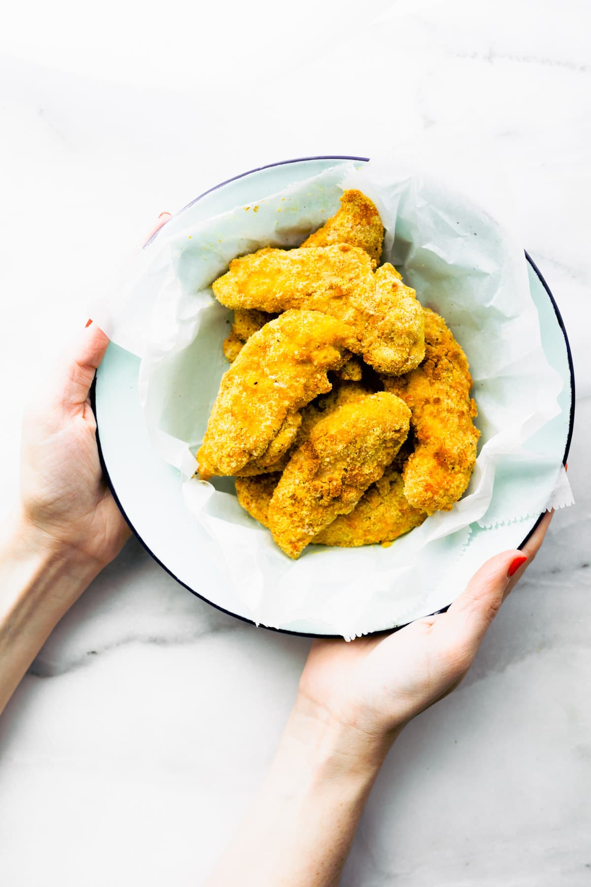 two hands holding white bowl full of crispy chicken strips