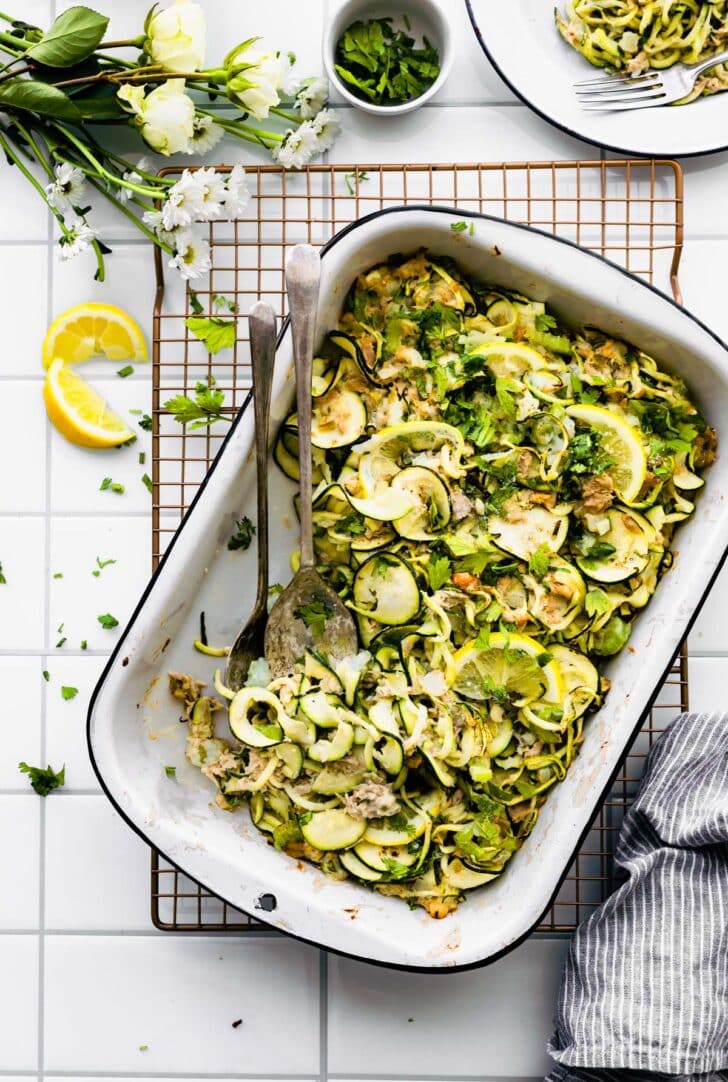 Paleo Tuna Green Chile Zoodle Casserole on white board with plate above and flowers, lemon, herbs on side