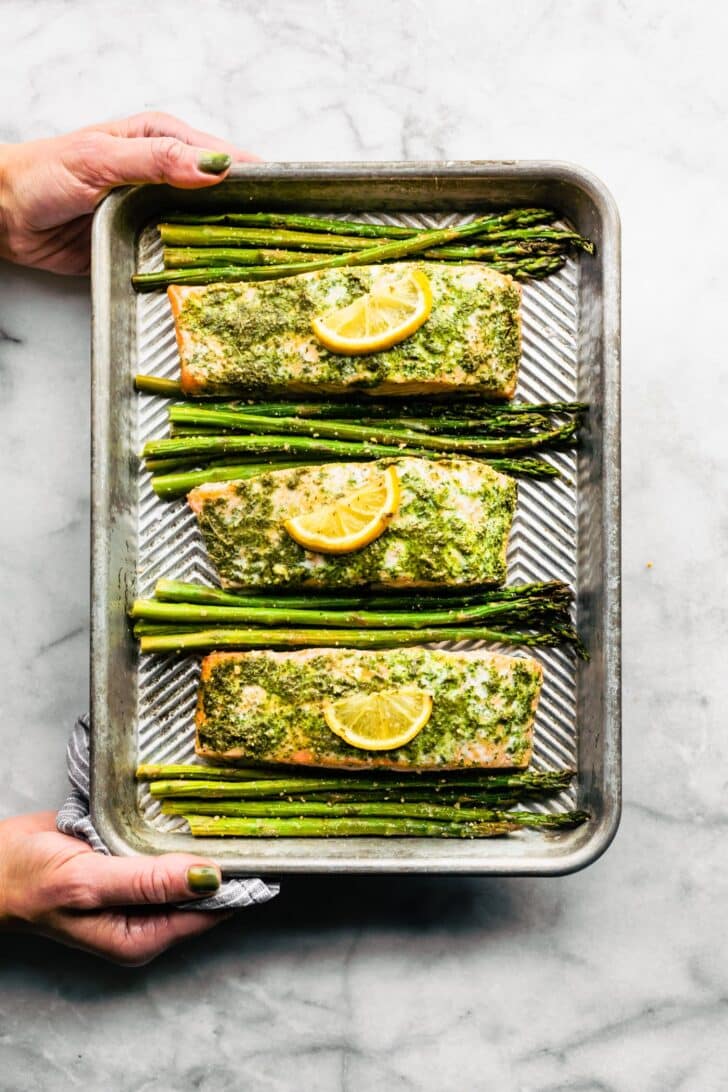 A woman's hands holding a metal sheet pan with three herb salmon filets and asparagus.