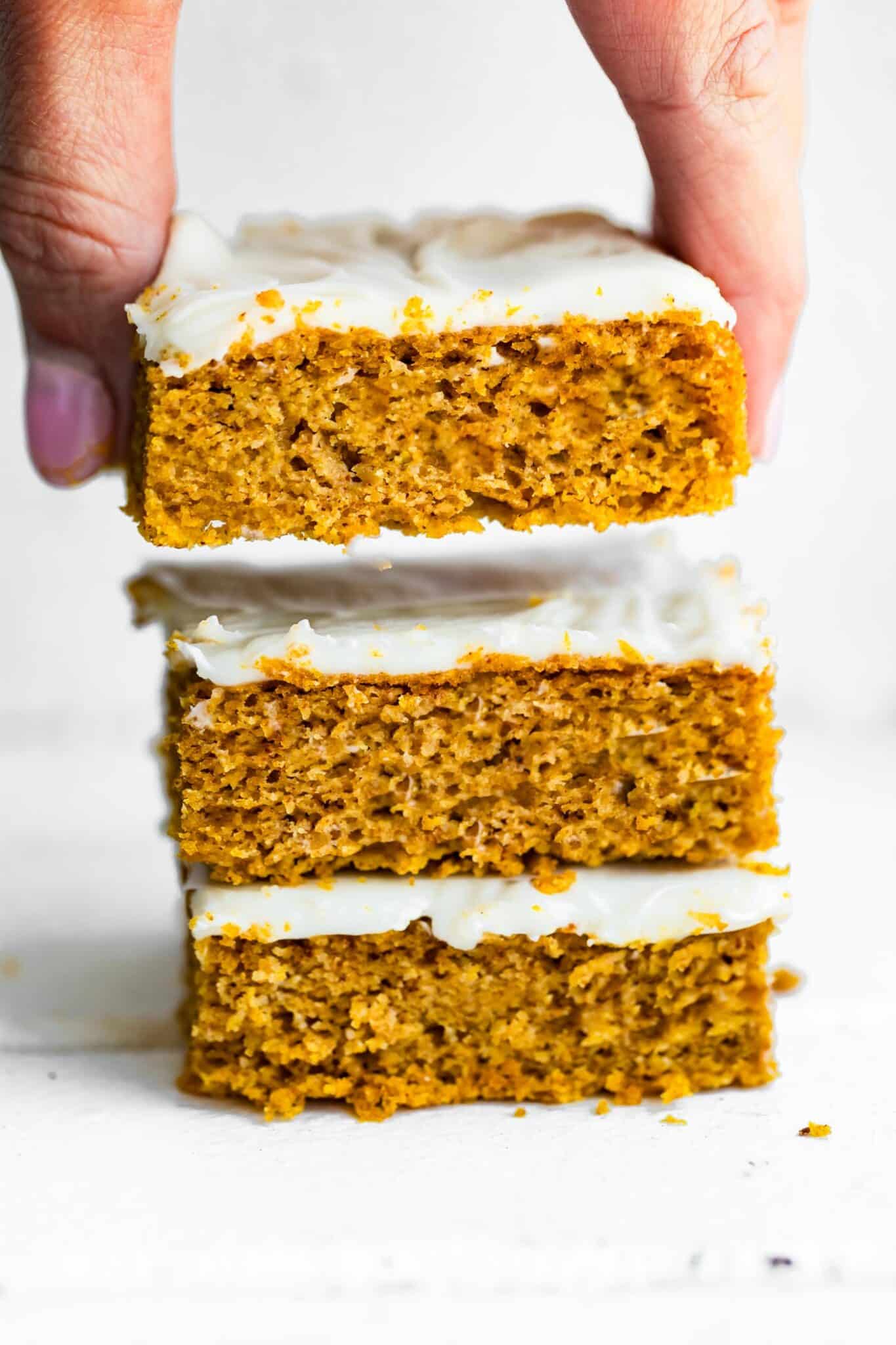 stack of frosted gluten free pumpkin bars with woman's hand grabbing the top bar