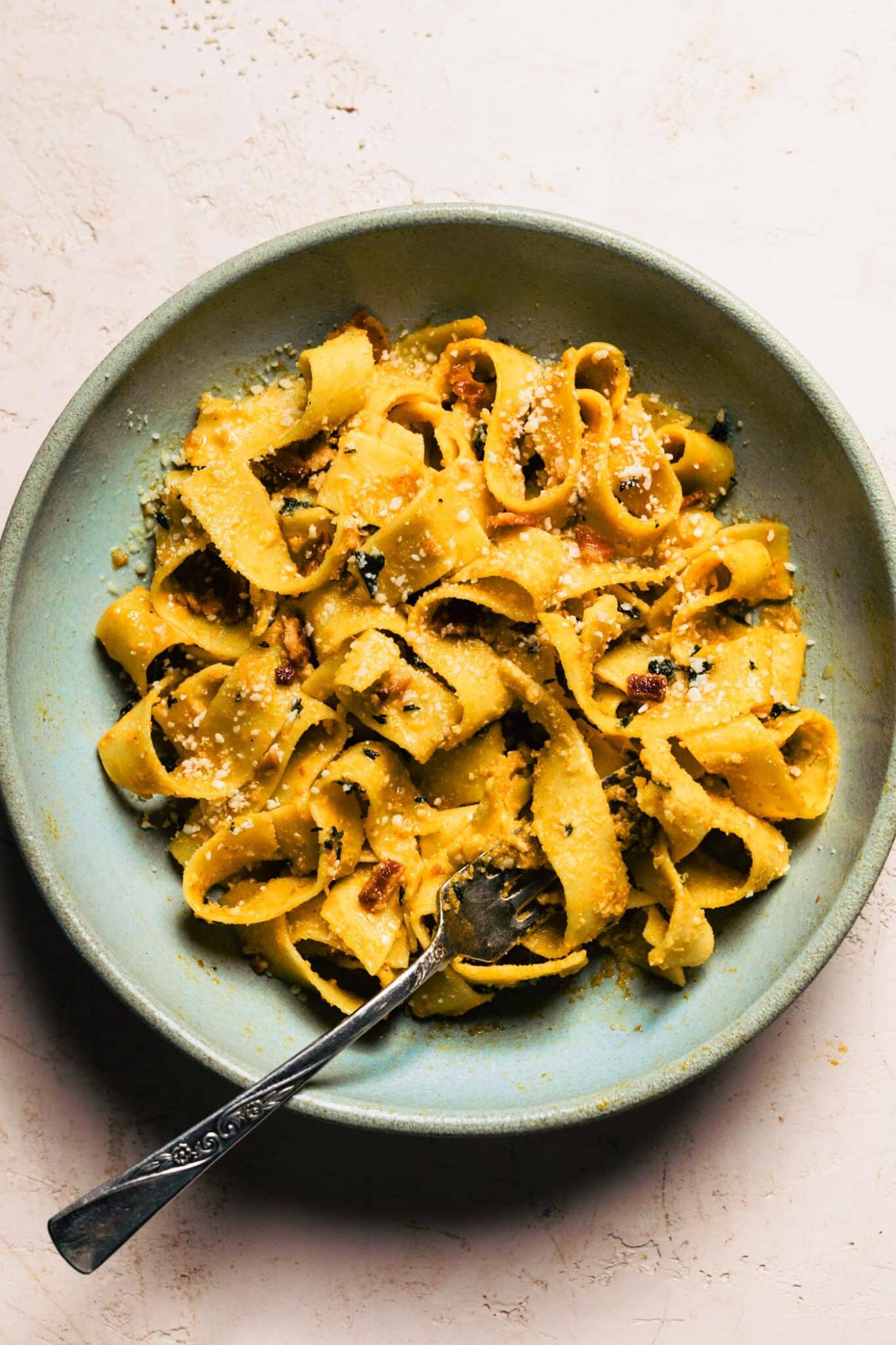 Close-up of fork twirling creamy pumpkin pappardelle pasta with crispy bacon pieces and grated parmesan in a blue ceramic bowl