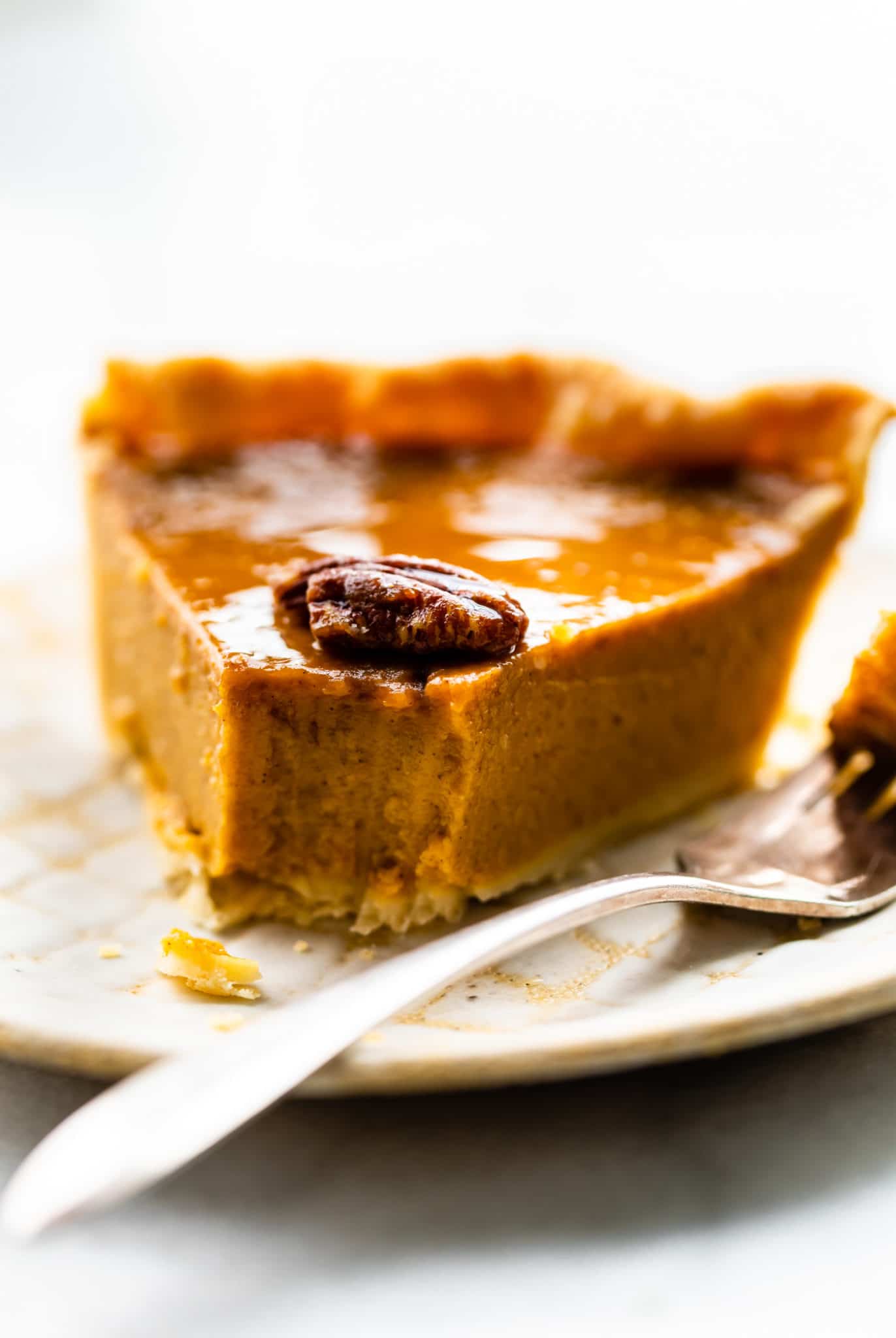 A slice of pumpkin pie on a plate front angle with tip of a piece taken off by fork. toasted pecan on top of pumpkin pie piece.