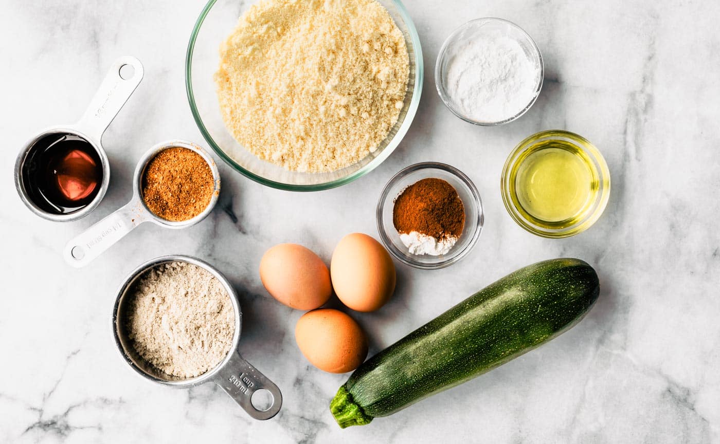 Ingredients arranged for making zucchini bread: bowl of flour, three eggs, bowl of spices and oil, fresh zucchini, and sugar in a measuring cup.