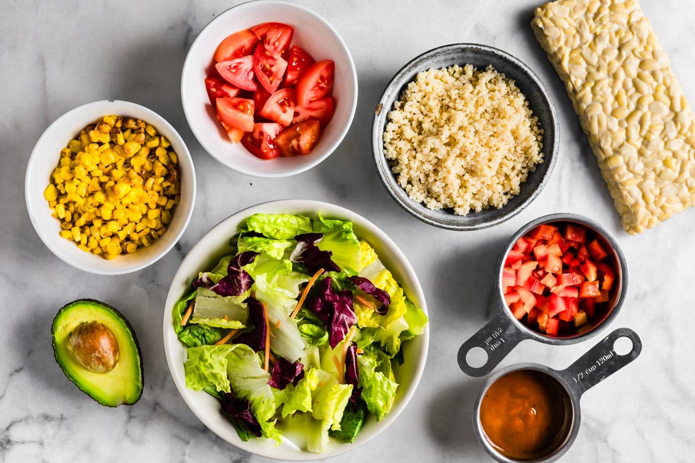 Ingredients in bowls for BBQ tempeh salad with quinoa on a white marble countertop.