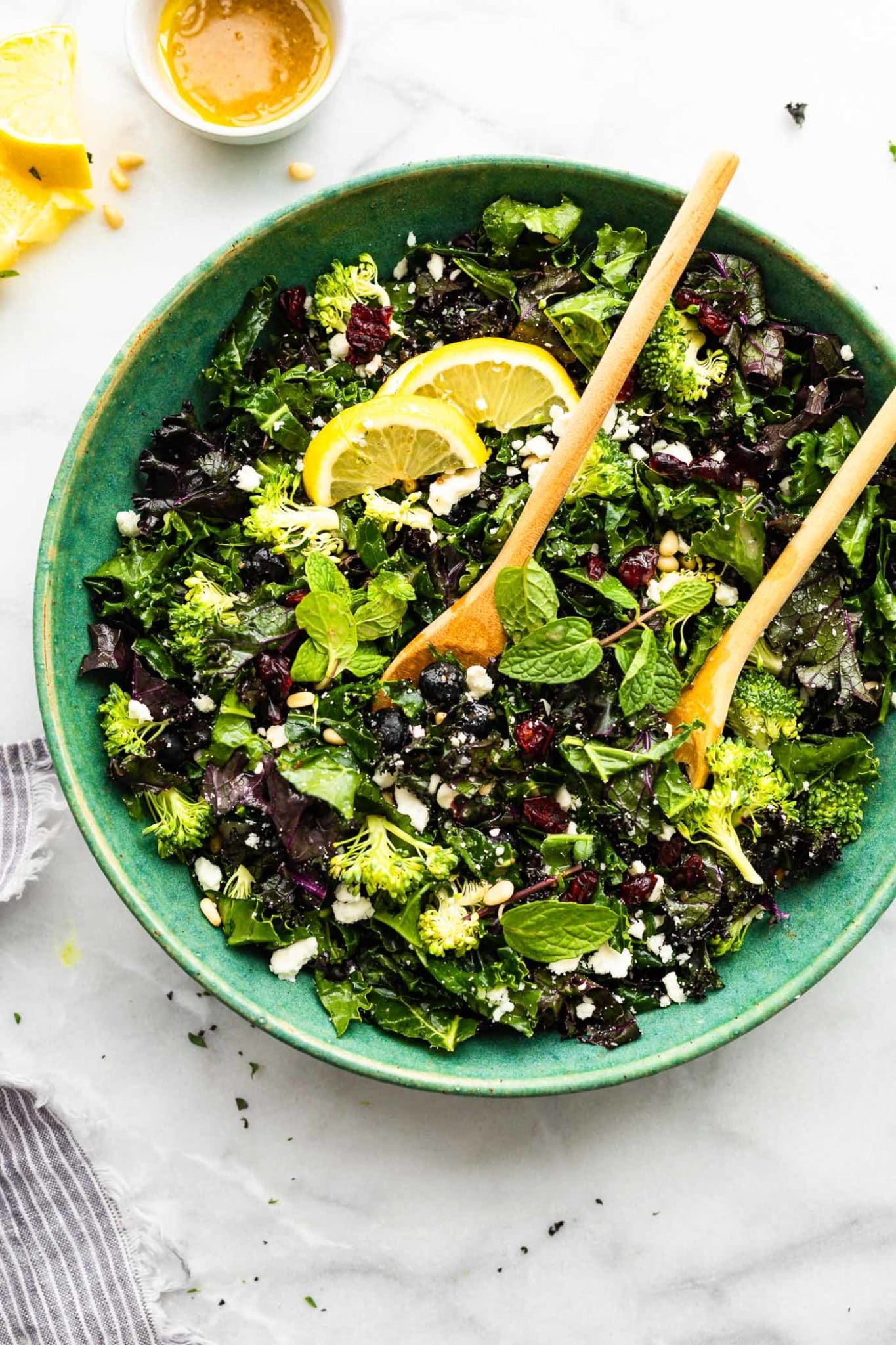 a large blue bowl filled with massaged kale salad topped with lemon wedges and two wooden serving spoons