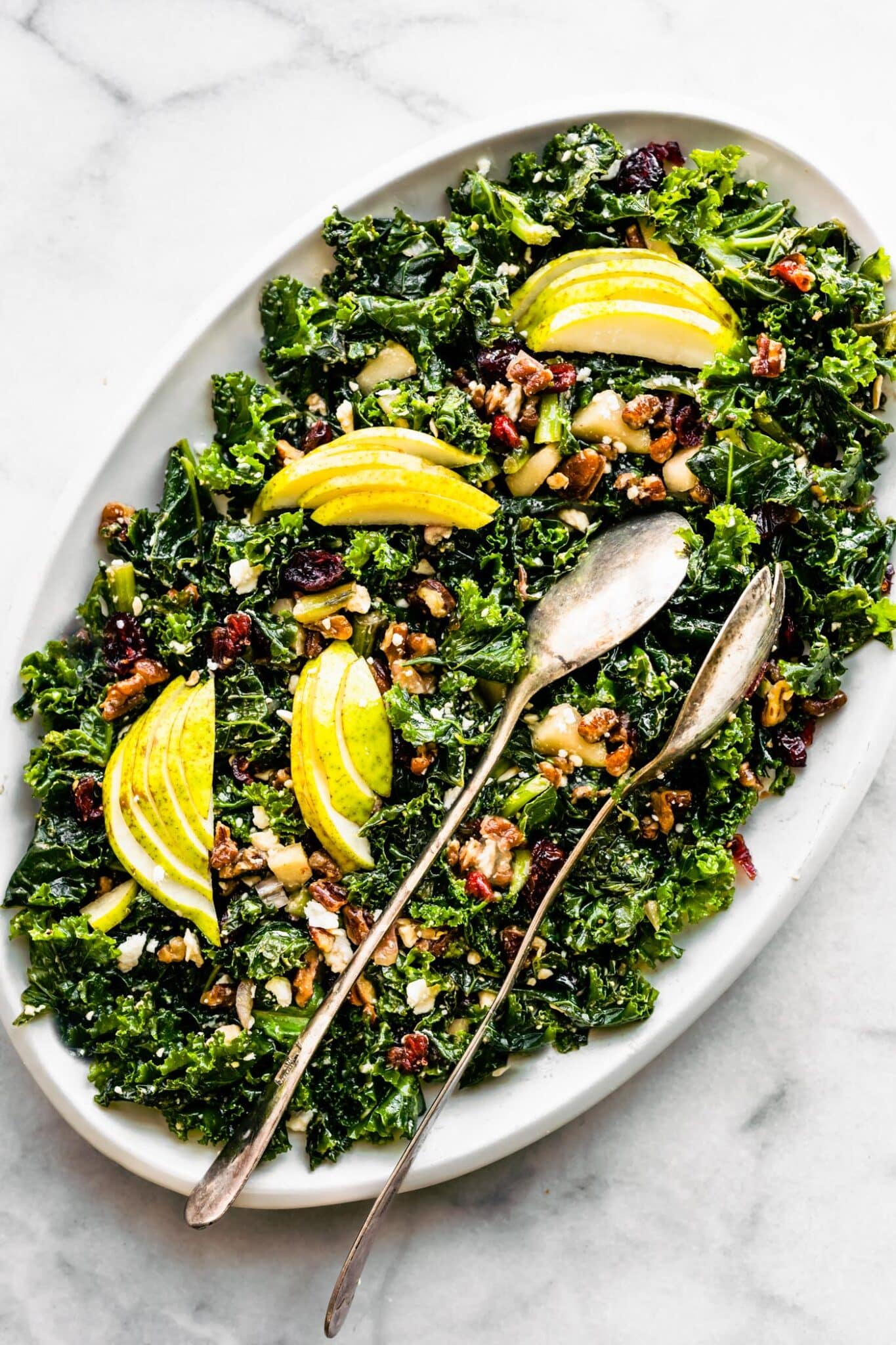 A white oval plate of goat cheese and pear kale salad with two serving spoons.