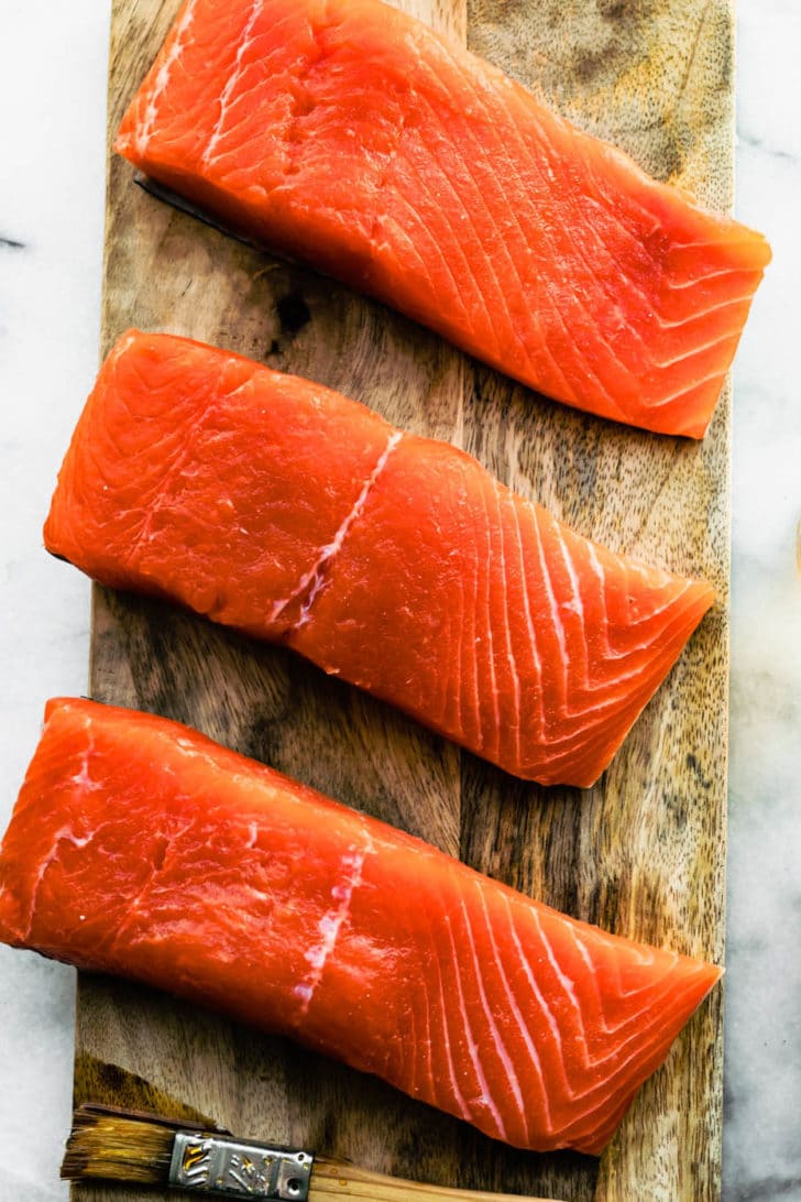 three pieces of raw salmon on a wooden plank with glaze ingredients on the side