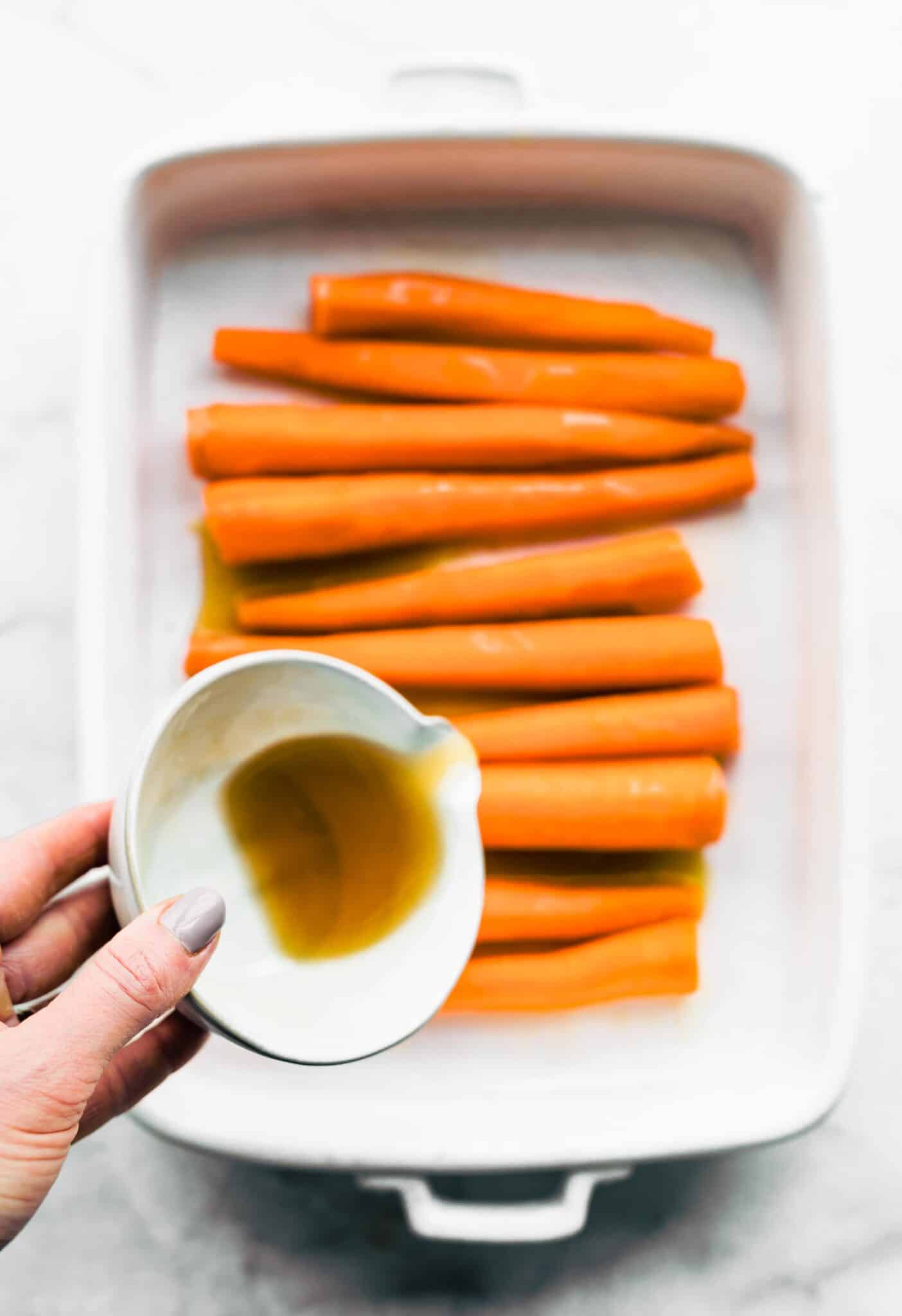 A woman's hand pouring a maple bourbon sauce over carrots in a white baking dish.