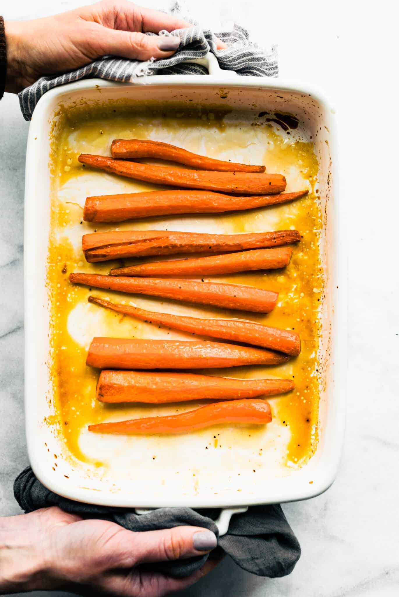 A woman's hand holding a hot pan of white baking dish with maple glazed carrots.