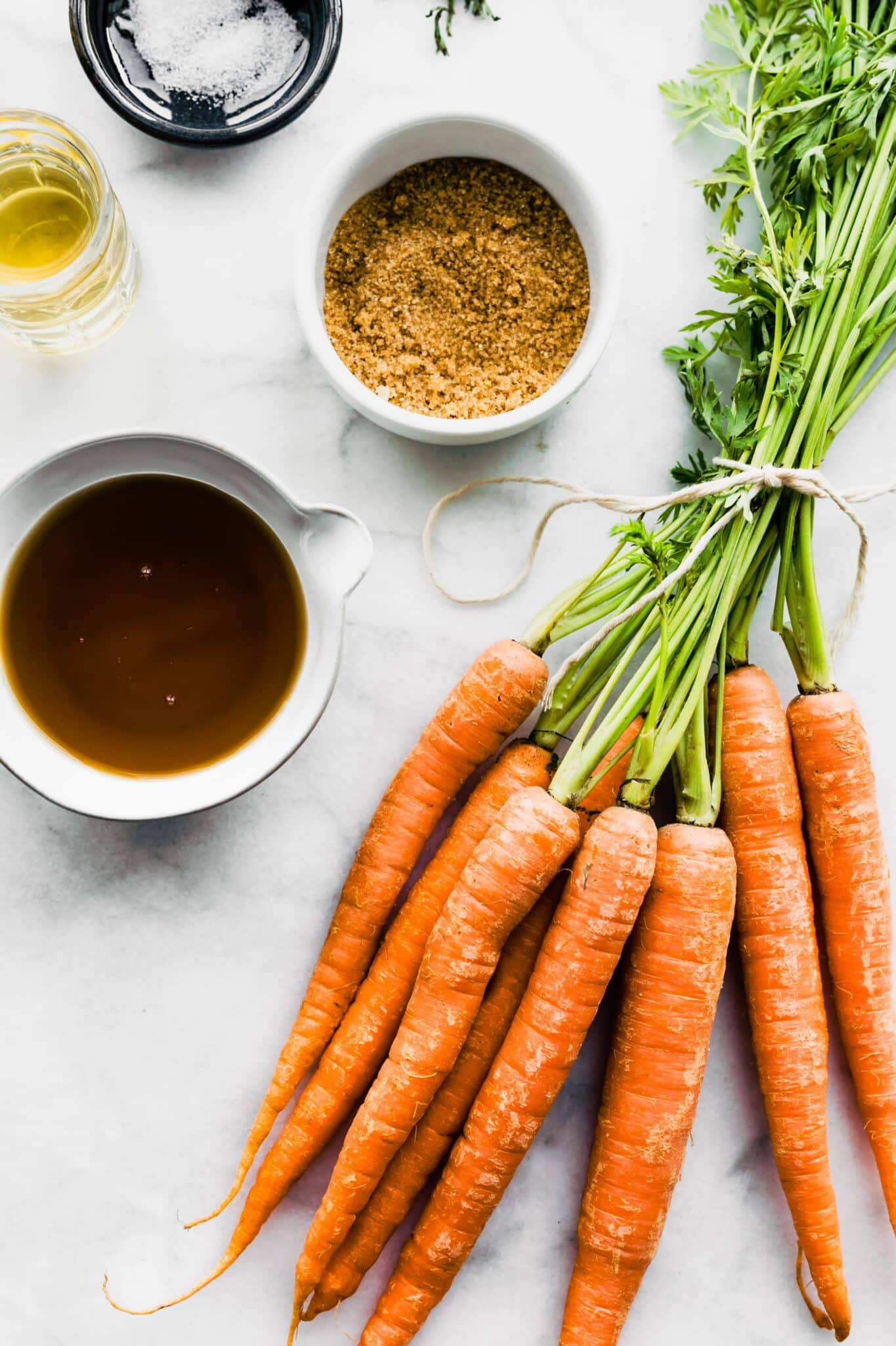 Raw carrots, maple syrup, oil, and salt in bowls on a white marble countertop.