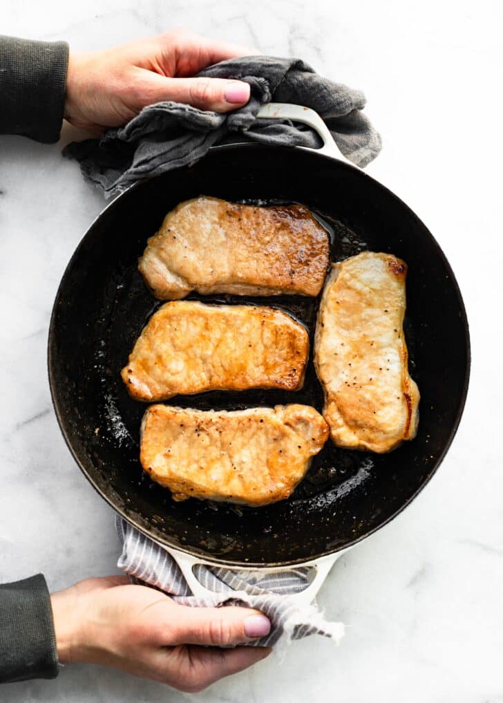 A woman holding a pan of four glazed pork chops in a cast iron pan.