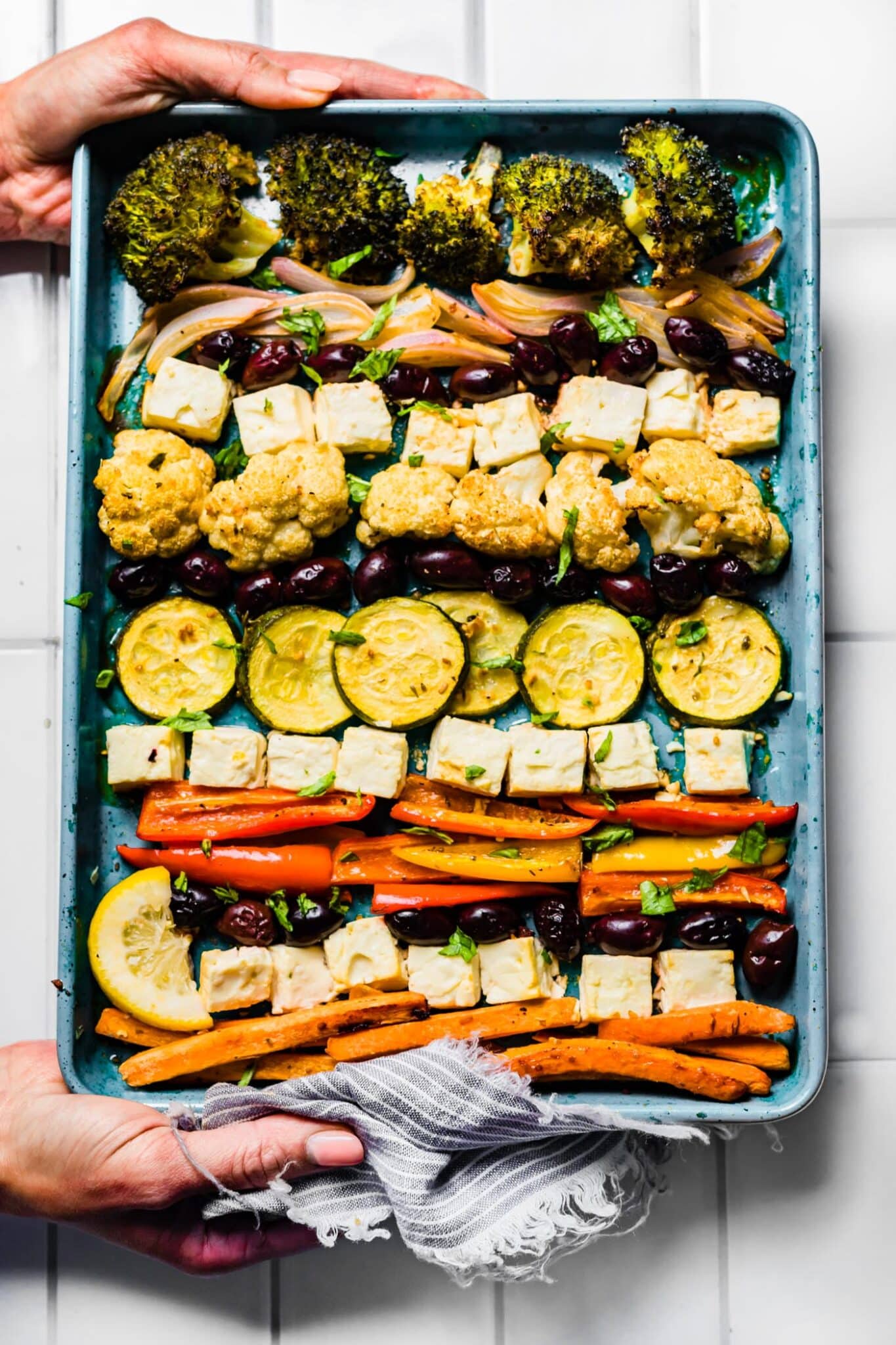 A woman's hands holding a blue sheet pan with Mediterranean roasted vegetables and feta.
