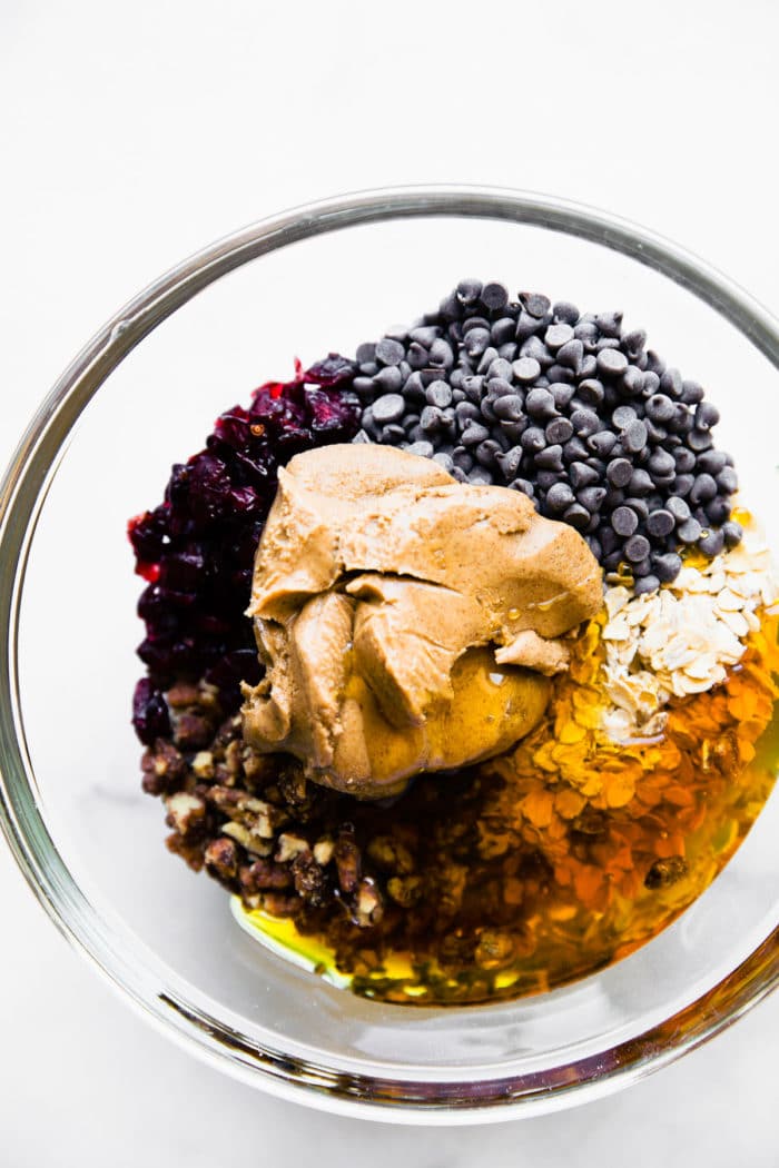 clear mixing bowl of chocolate chips, nut butter, cranberries, oats, nuts and honey. View from above