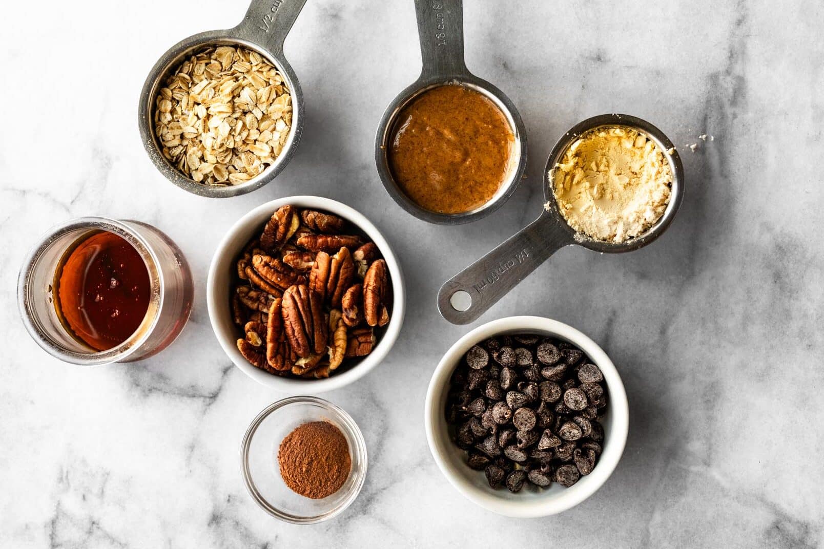 Overhead view of pecan pie ball ingredients arranged on a marble surface, including oats, pecans, chocolate chips, cinnamon, almond butter, protein powder, and maple syrup