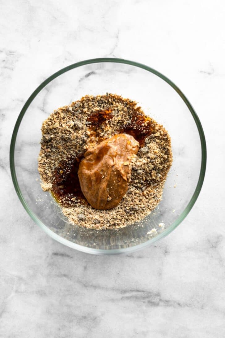 Glass bowl with ground oat-pecan mixture topped with almond butter and maple syrup before mixing