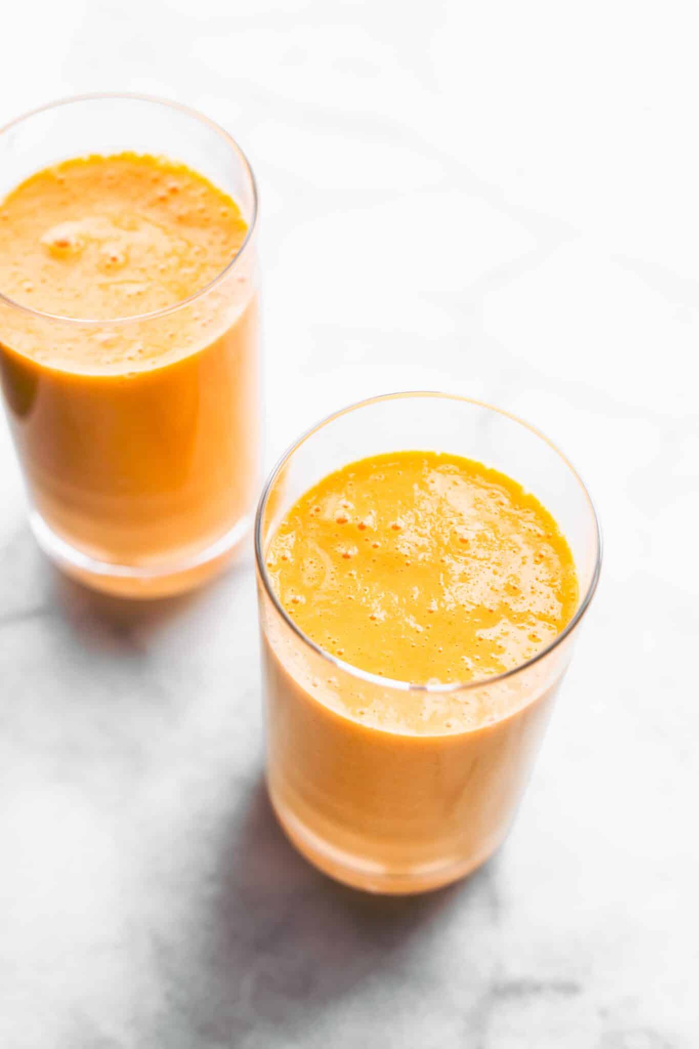 Two glasses of orange carrot smoothie in glasses on a white marble countertop.
