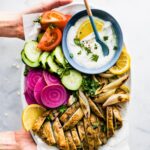 A woman's hands holding a white platter of oven roasted chicken shawarma and shawarma sauce.