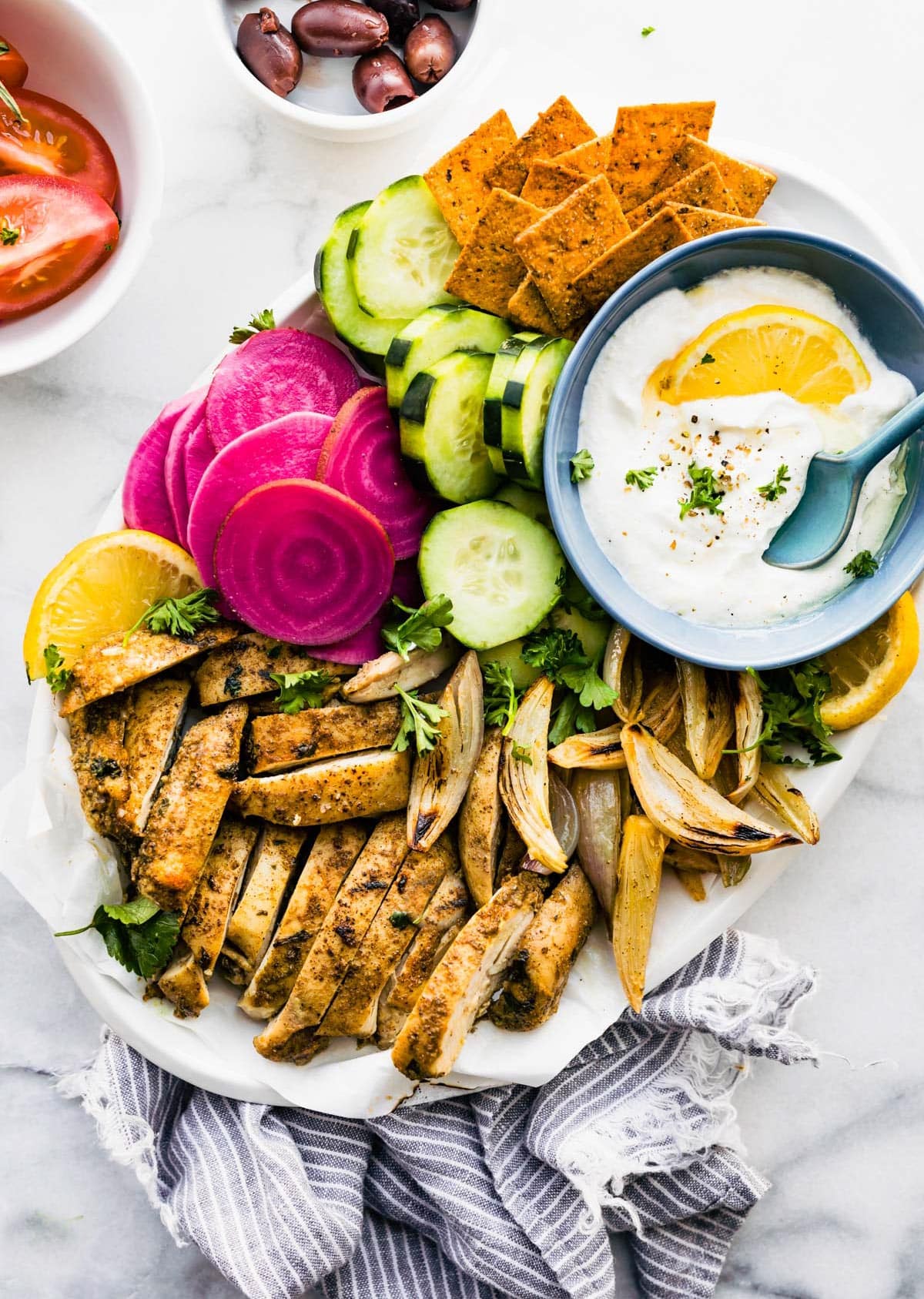 A white platter of oven roasted chicken shawarma, fresh vegetables, crackers and shawarma sauce.