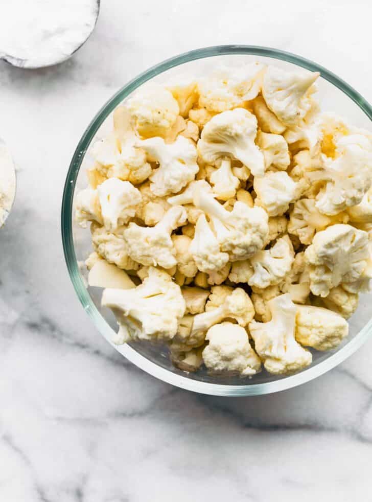 Cauliflower florets in a glass bowl on a white marble countertop.