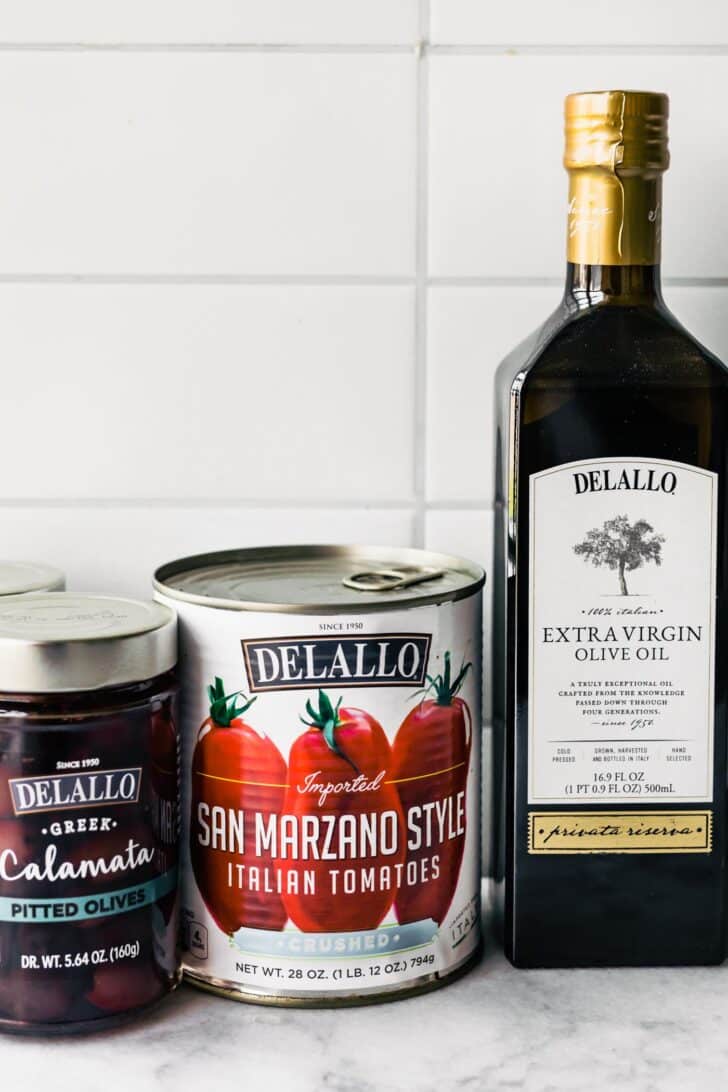 Delallo foods jarred Calamata olives, canned San Marzano tomatoes and olive oil on a countertop.