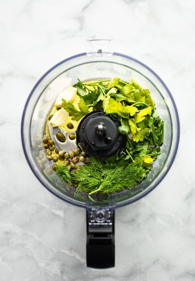 Food processor filled with fresh herbs, lemon juice, oil and capers before blending.