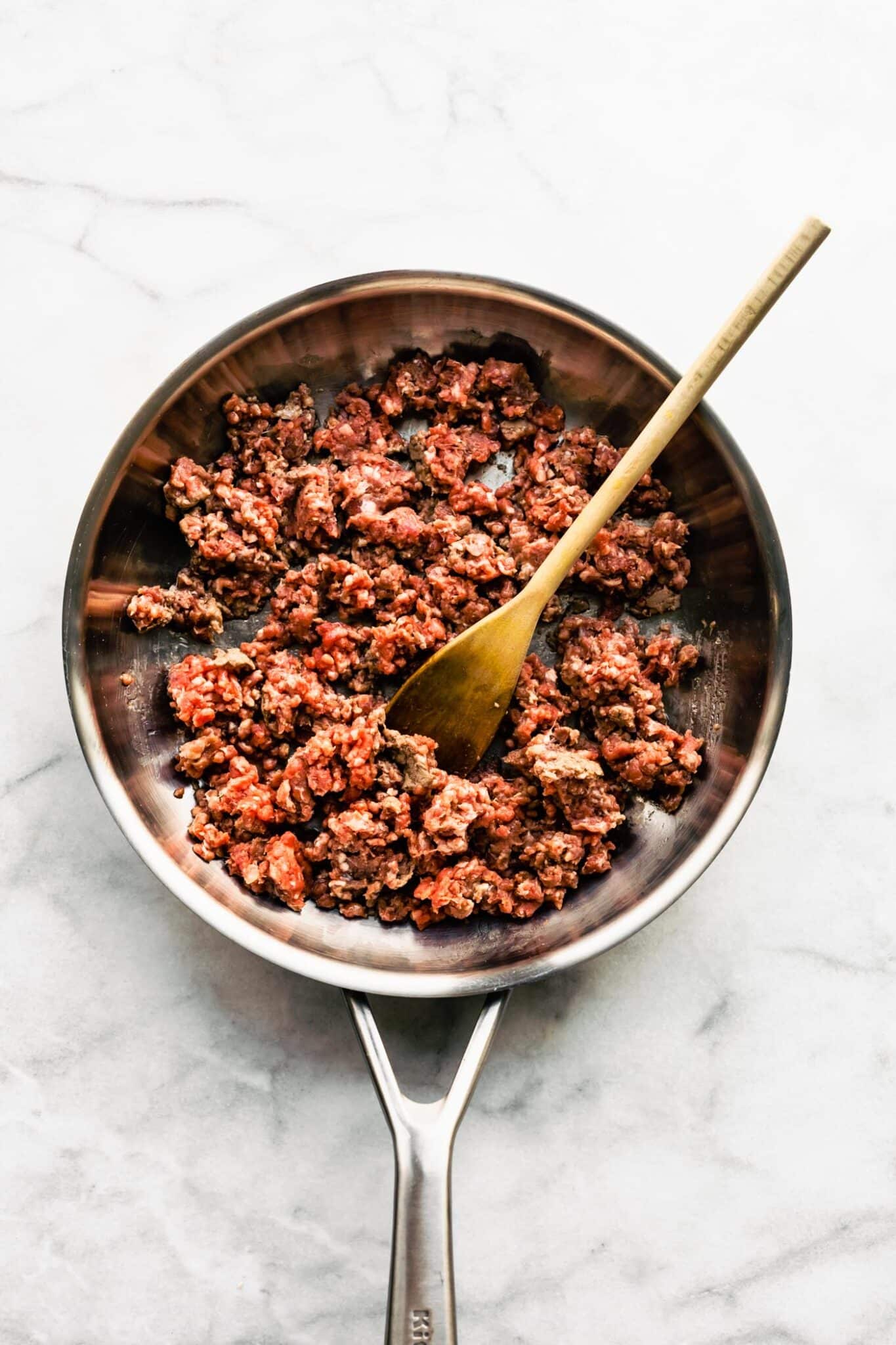 A wooden spoon in a stovetop pan of ground beef ready to brown.