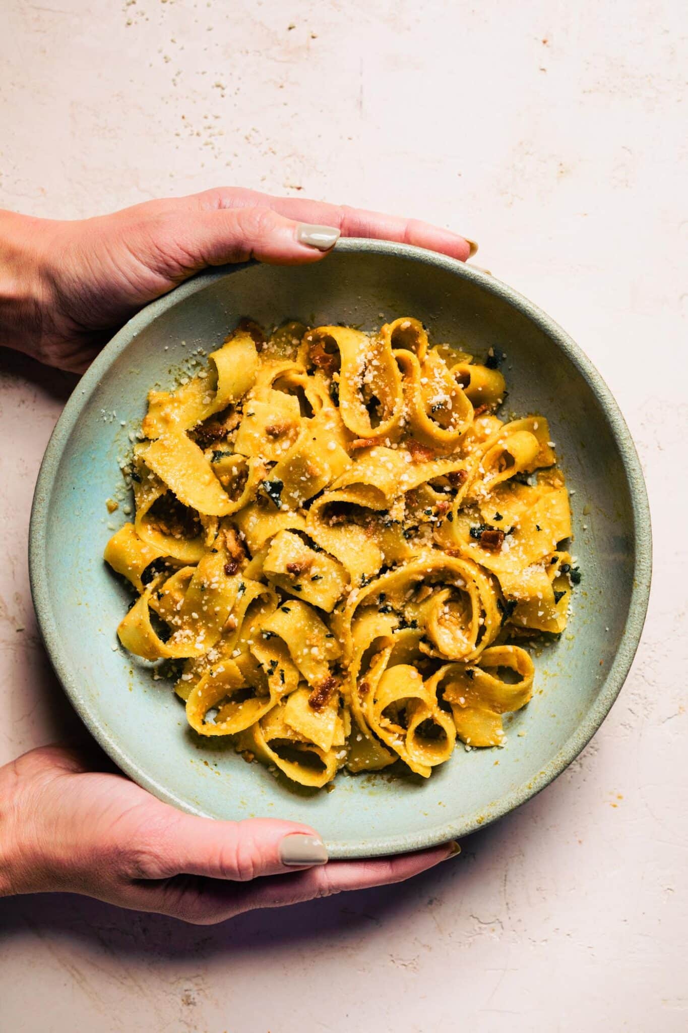 Hands holding bowl of gluten-free pumpkin pasta with bacon and parmesan on light background