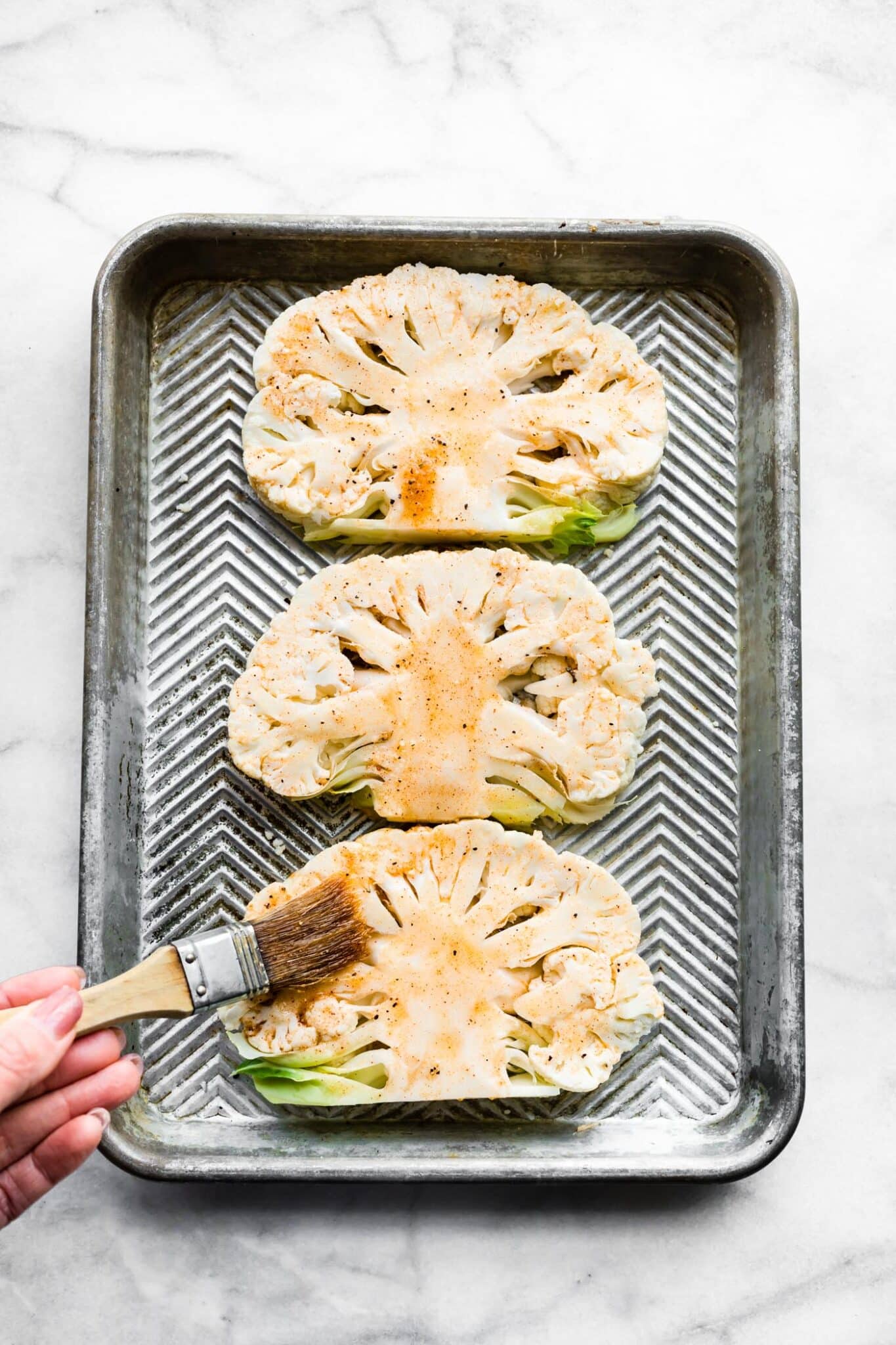 Three cauliflower steaks on a sheet pan with a woman brushing marinade on them.