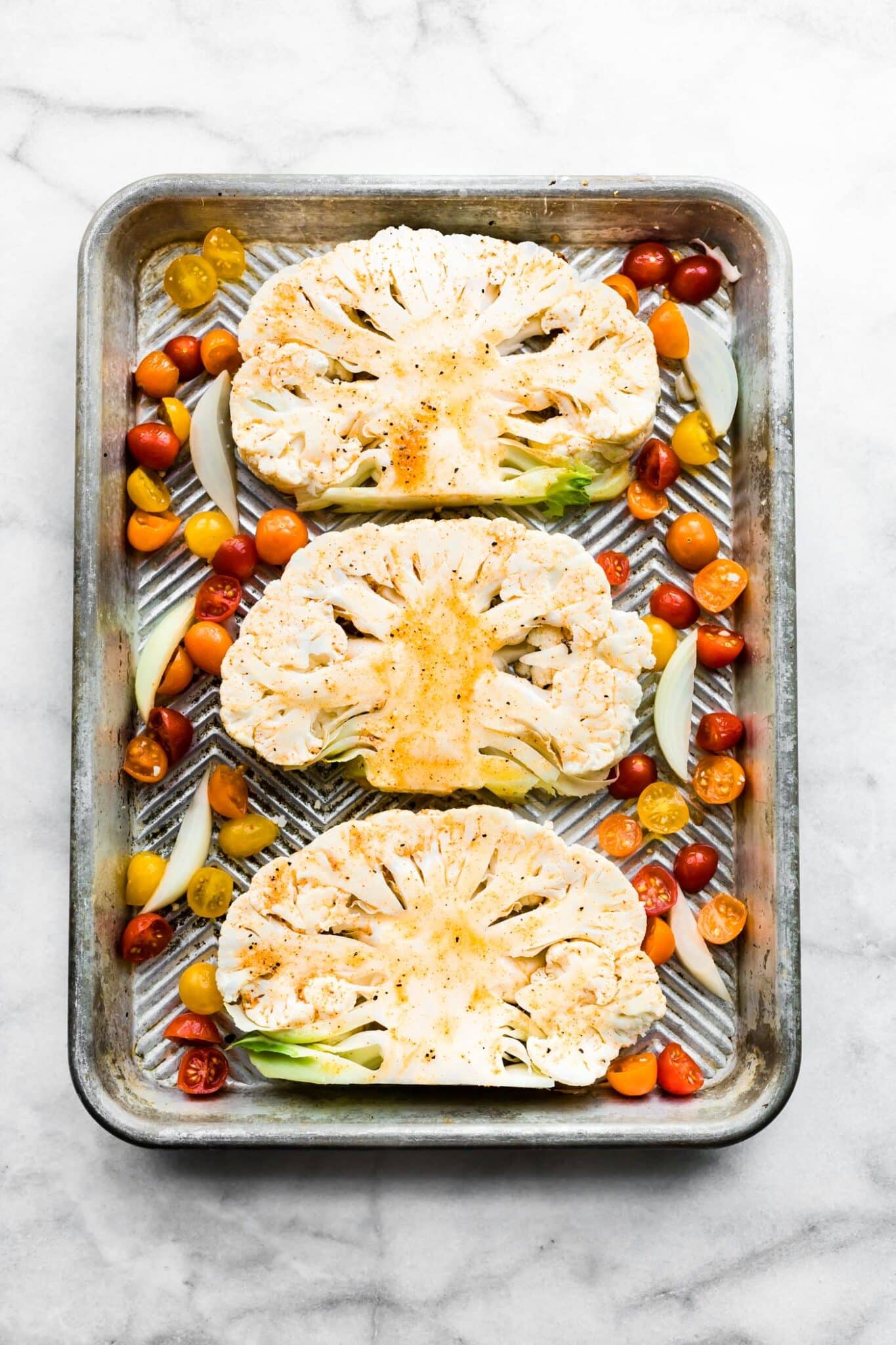 Three raw seasoned cauliflower steaks on a sheet pan with tomatoes and onion.