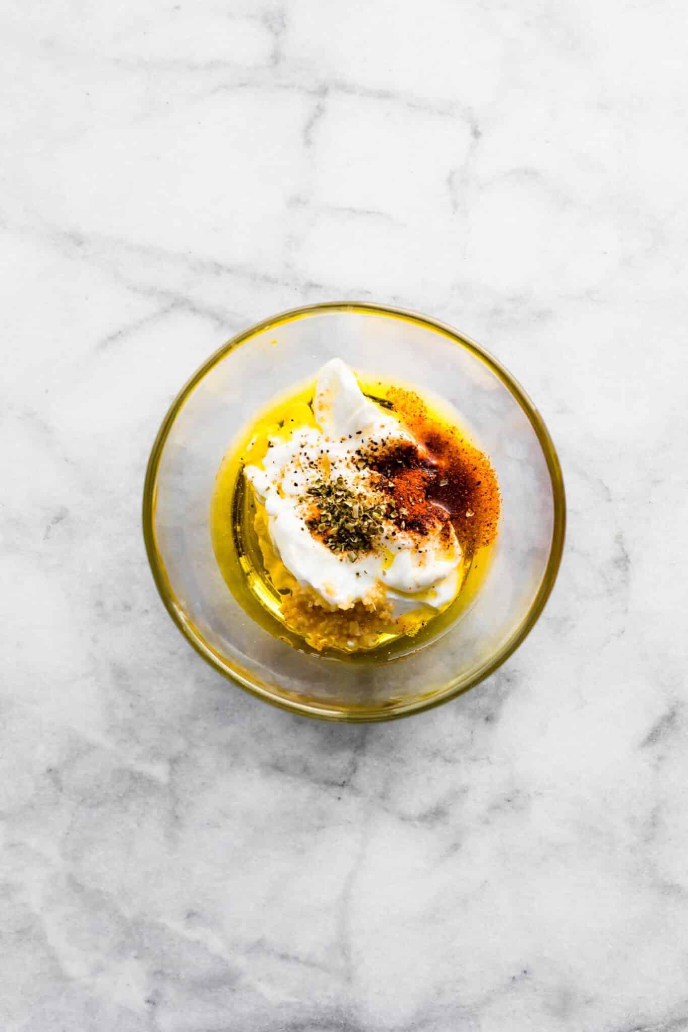Yogurt, olive oil, and spices in a clear bowl on a white marble countertop.