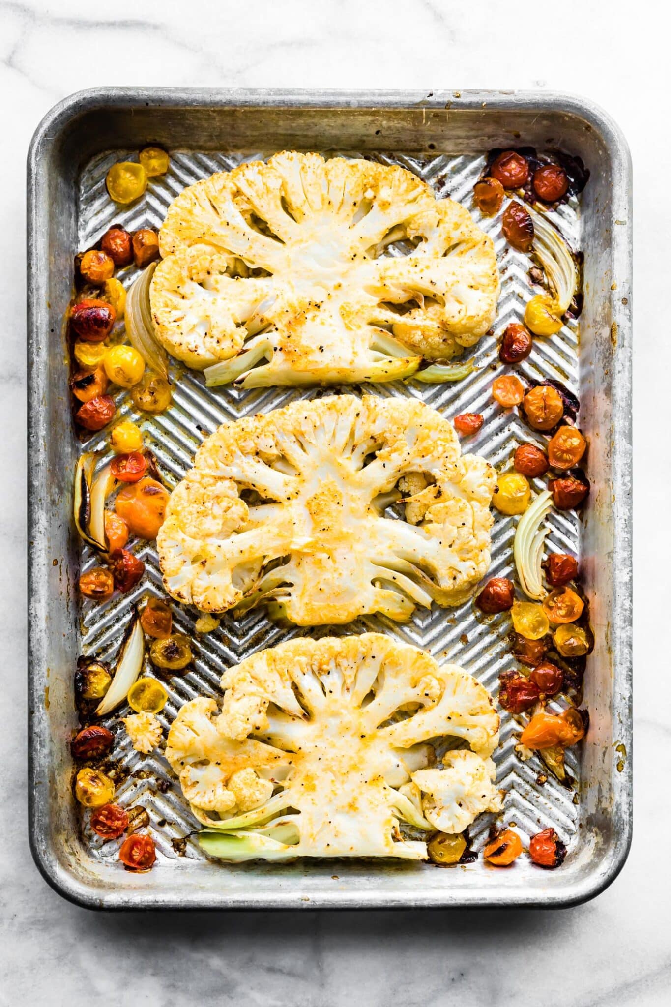 Three baked cauliflower steaks on a sheet pan with tomatoes and sliced onion.
