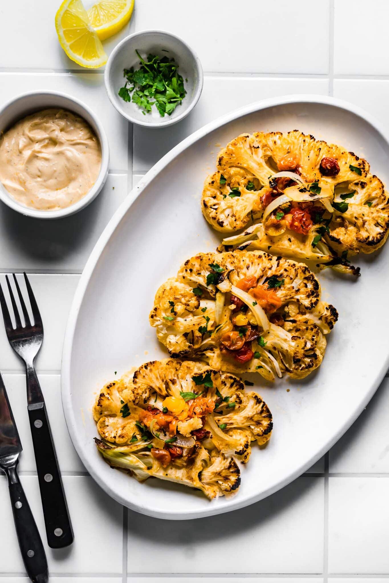 Three roasted cauliflower steaks on a white plate with blistered tomatoes and chopped herbs.