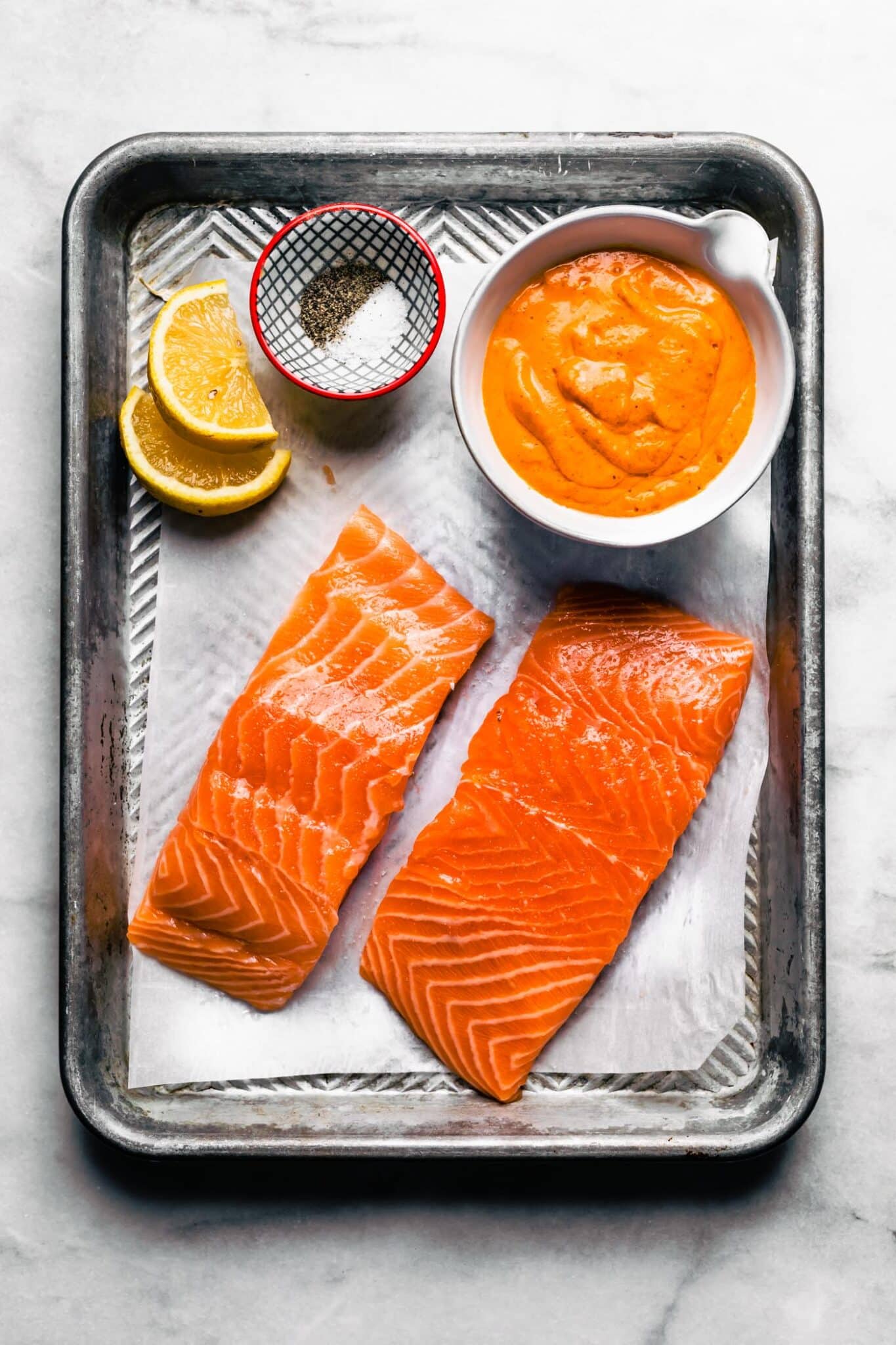 sheet pan of ingredients for red pepper salmon. Bowl of roasted red pepper sauce, salmon fillets, lemon, spices