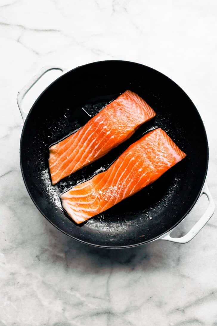 2 raw salmon fillets in a black round pan with oil and seasoning