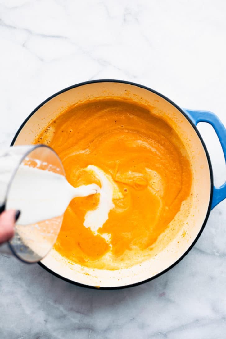 a pot of roasted root vegetable soup with a woman's hand pouring a measuring cup filled with non-dairy milk into it