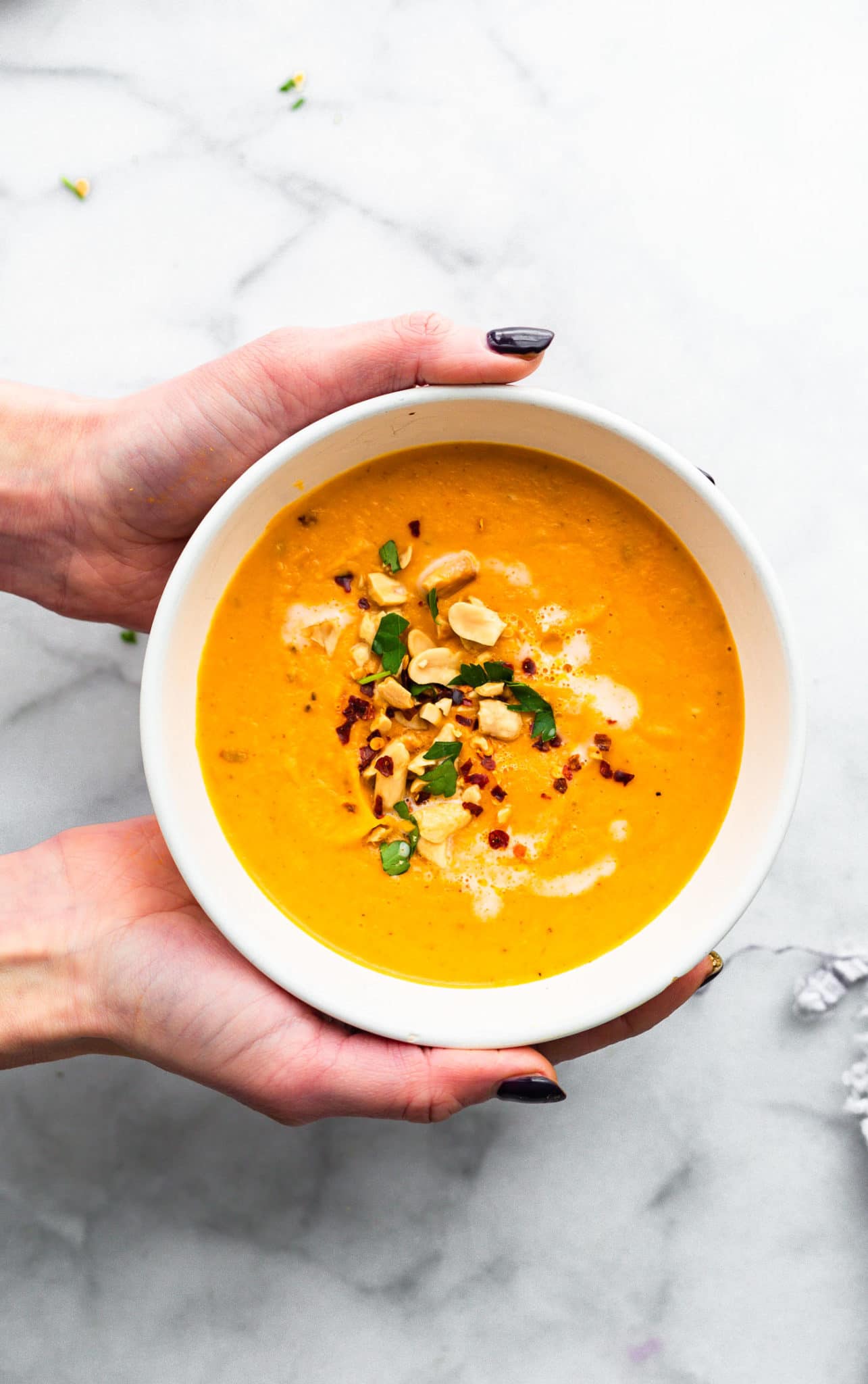 two hands holding a white bowl of roasted root vegetable soup topped iwth crushed nuts, chili pepper flakes, and herbs