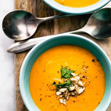 a blue bowl of root vegetable soup topped with curshed nuts and herbs with two spoons on the side