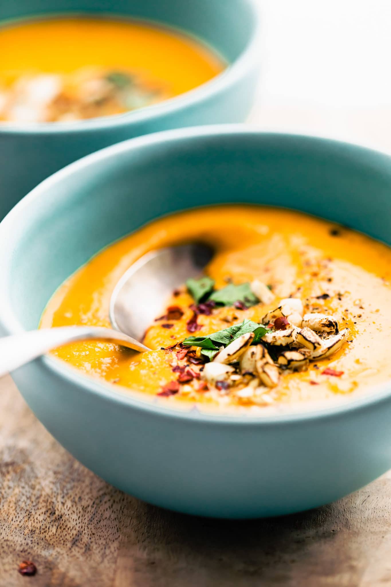 a closeup side shot of cumin roasted root vegetable soup in a blue bowl wopped with red pepper flakes, crushed nuts, and fresh herbs with a spoon in it