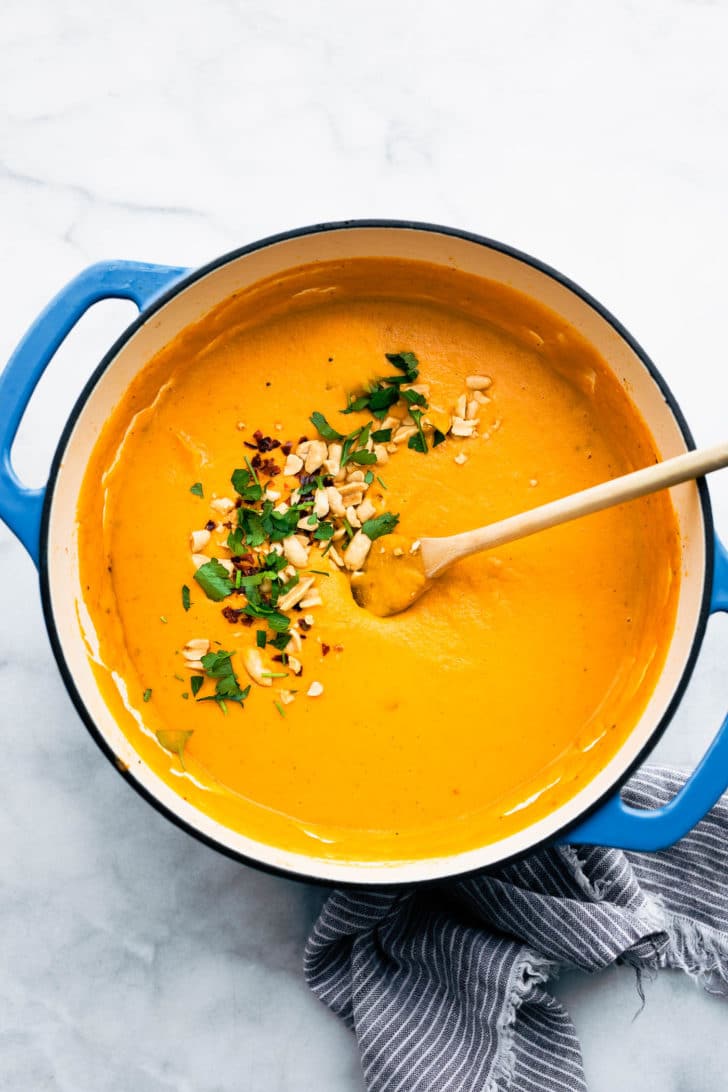 a pot of roasted root vegetable soup topped with crushed nuts, red pepper flakes, and herbs being stirred with a wooden spoon