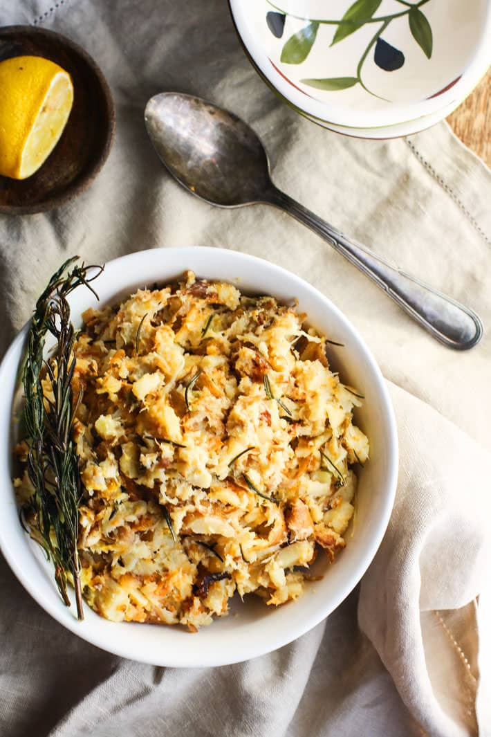 A bowl of rosemary carrot & parsnip mash sitting on a linen tablecloth with a spoon next to the bowl.