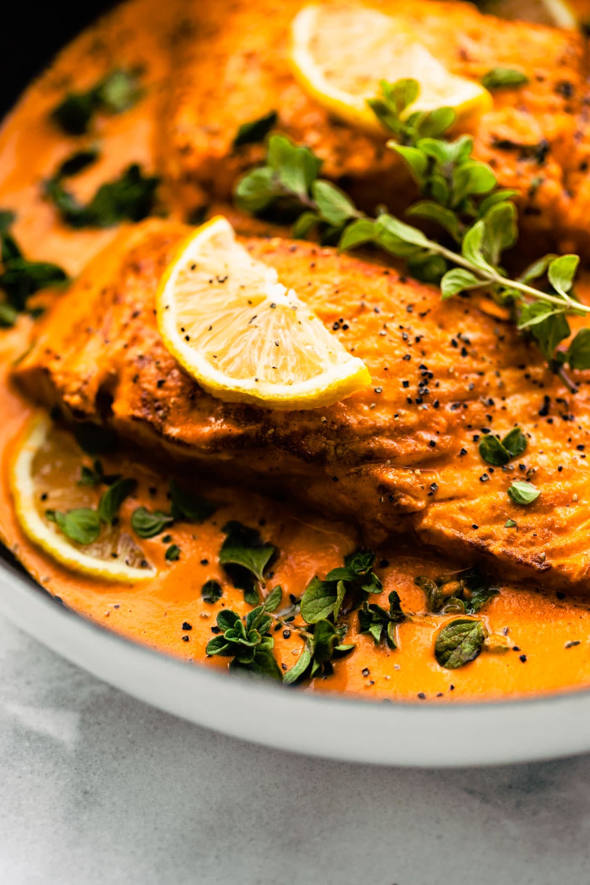Close up view baked salmon in roasted red pepper sauce with lemon slice on top with fresh oregano.