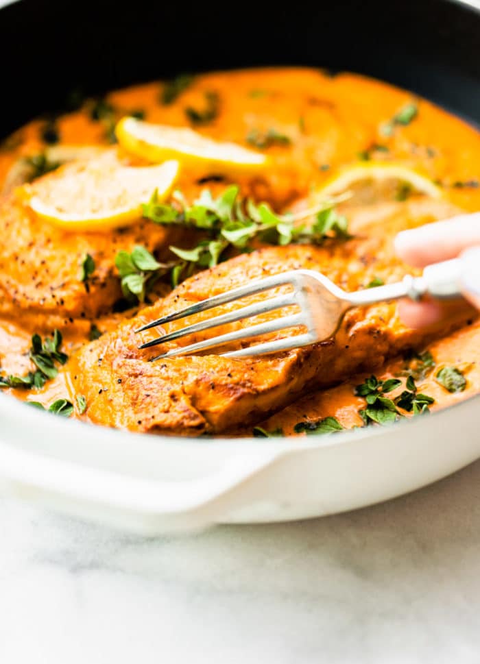 A fork cutting into baked salmon in red pepper sauce still in a skillet.