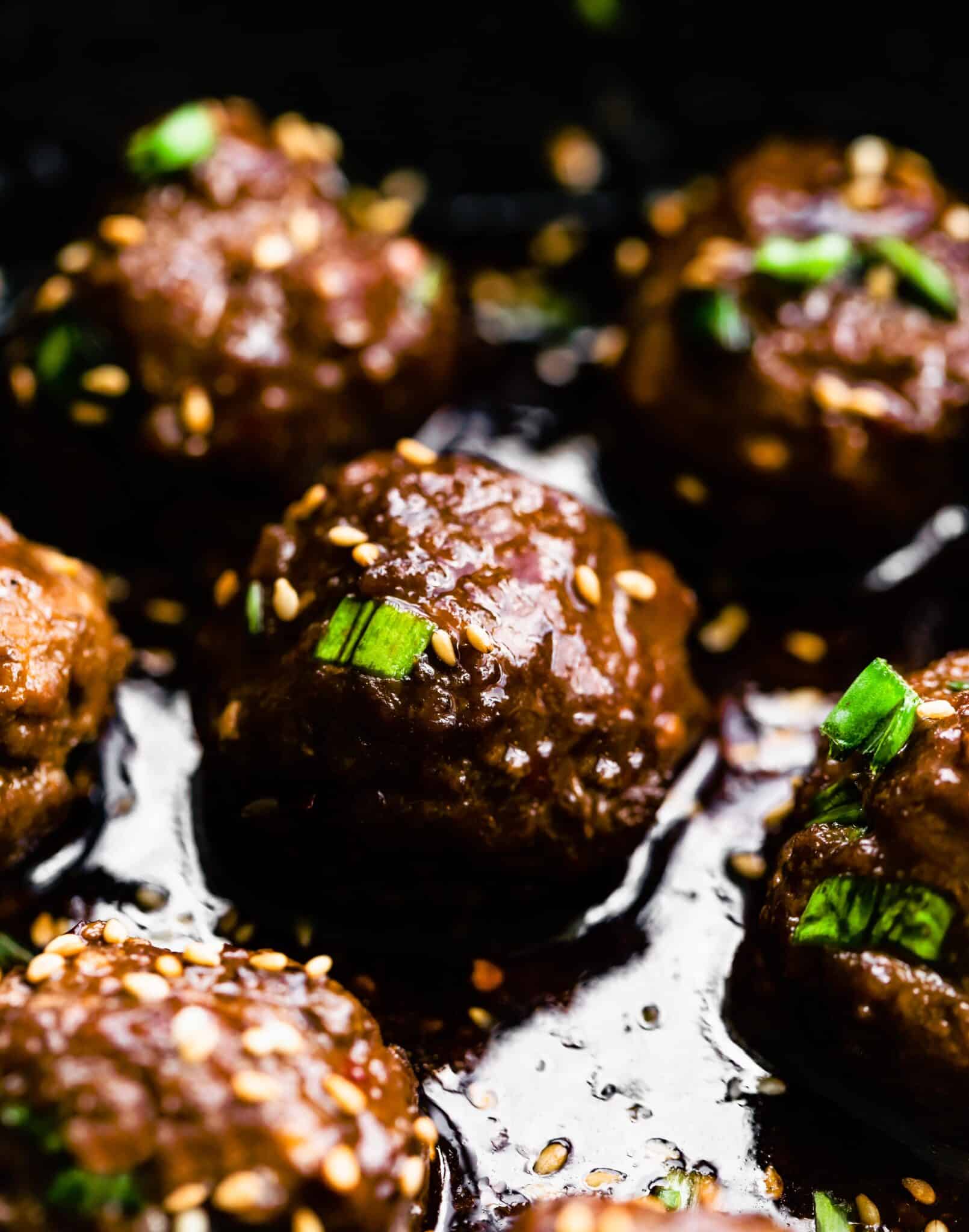 Up close photo of saucy Asian meatball topped with white sesame seeds and green onion.
