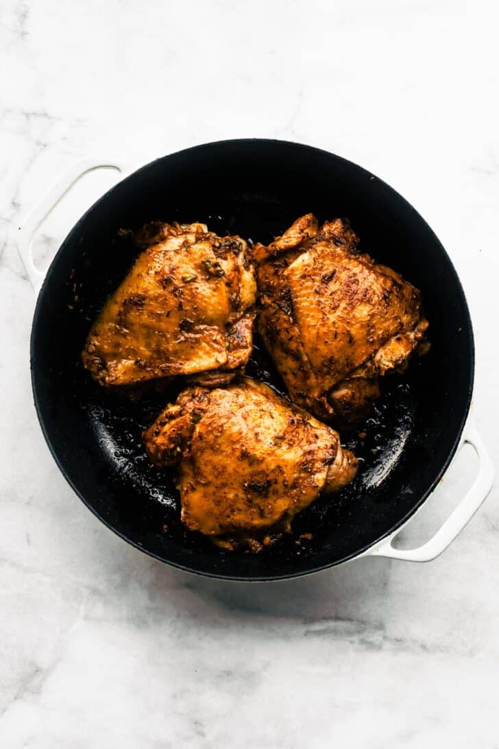 3 seared skin-on chicken thighs coated in jerk marinade, sitting in a black skillet