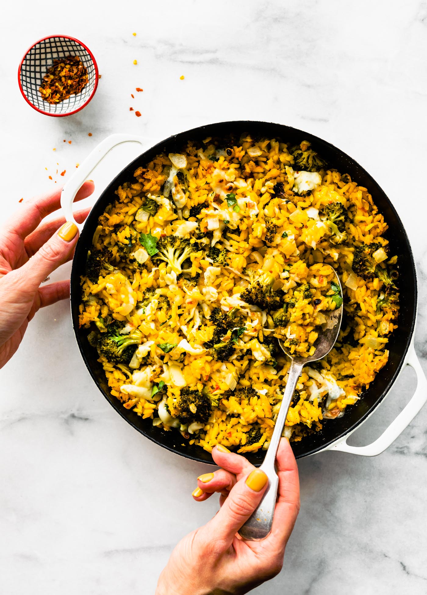 woman's hands scooping broccoli cheese casserole with gluten free orzo in a white round casserole dish