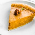 Slice of pumpkin pie on a white plate topped with candied nuts.